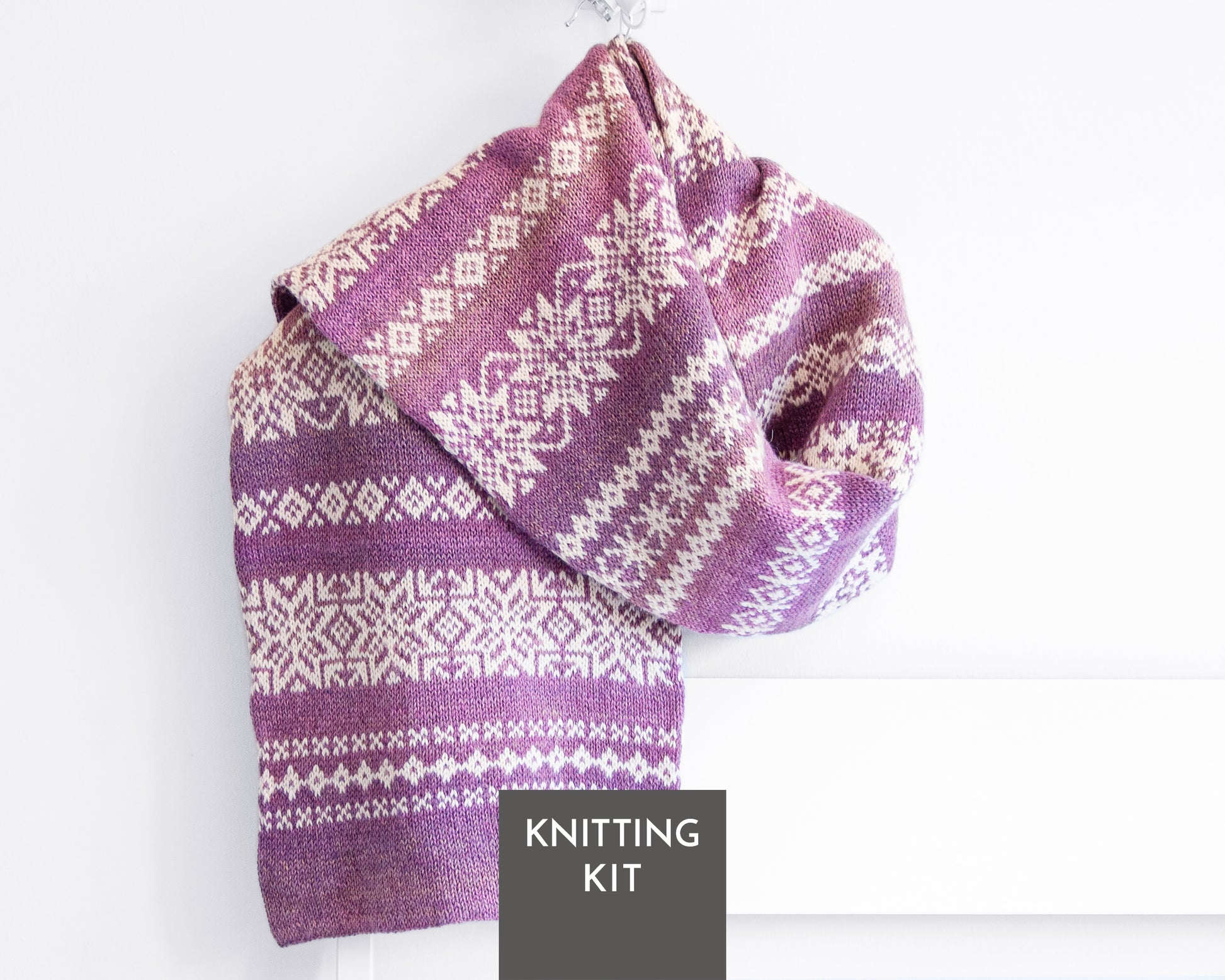 A folded purple and white Fair Isle knit scarf, hanging against a white wall. A dark gray label at the bottom reads 'KNITTING KIT'.