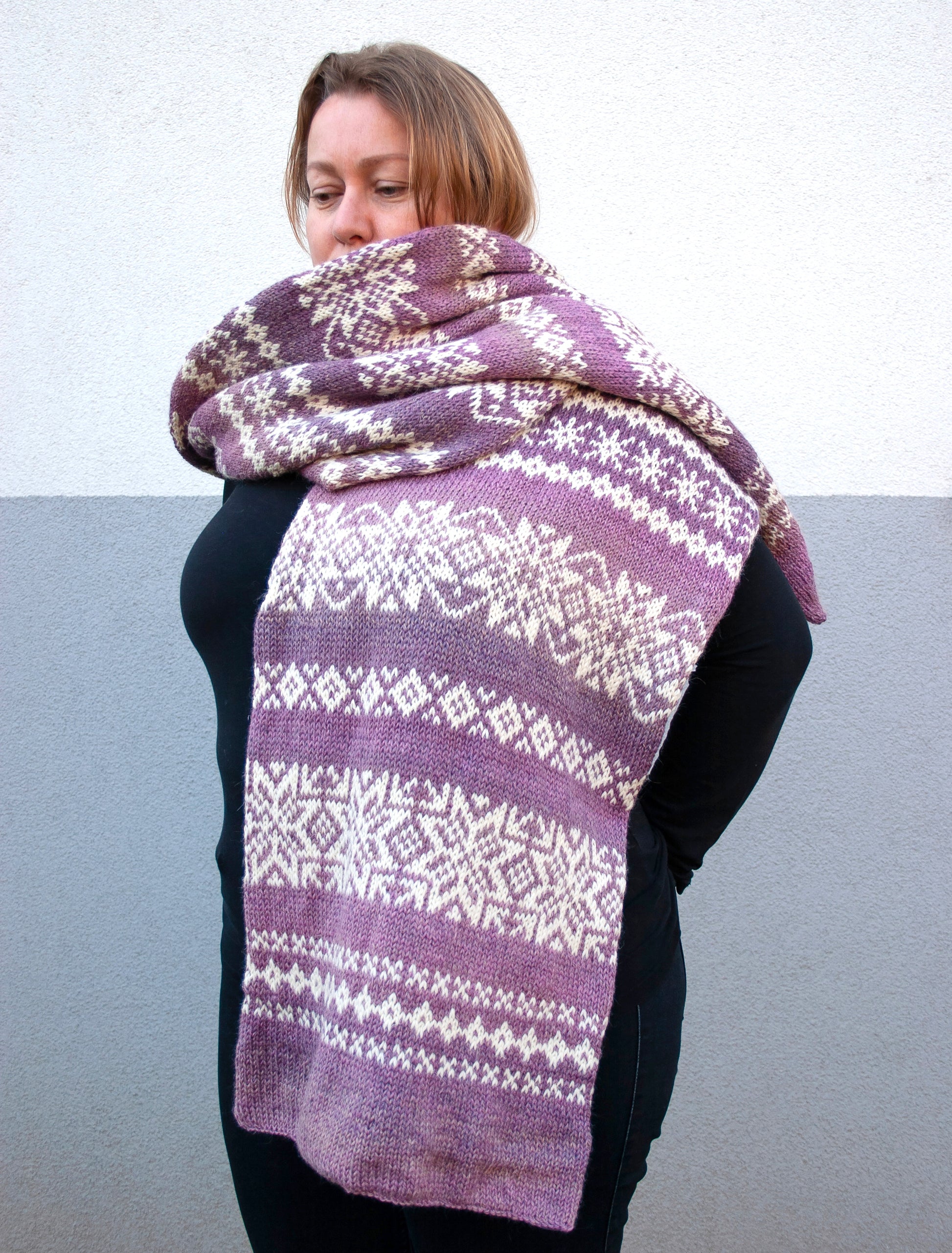 A woman wearing a purple and cream hand-knitted Fair Isle scarf with traditional Nordic snowflake and geometric patterns, wrapped around her shoulders.