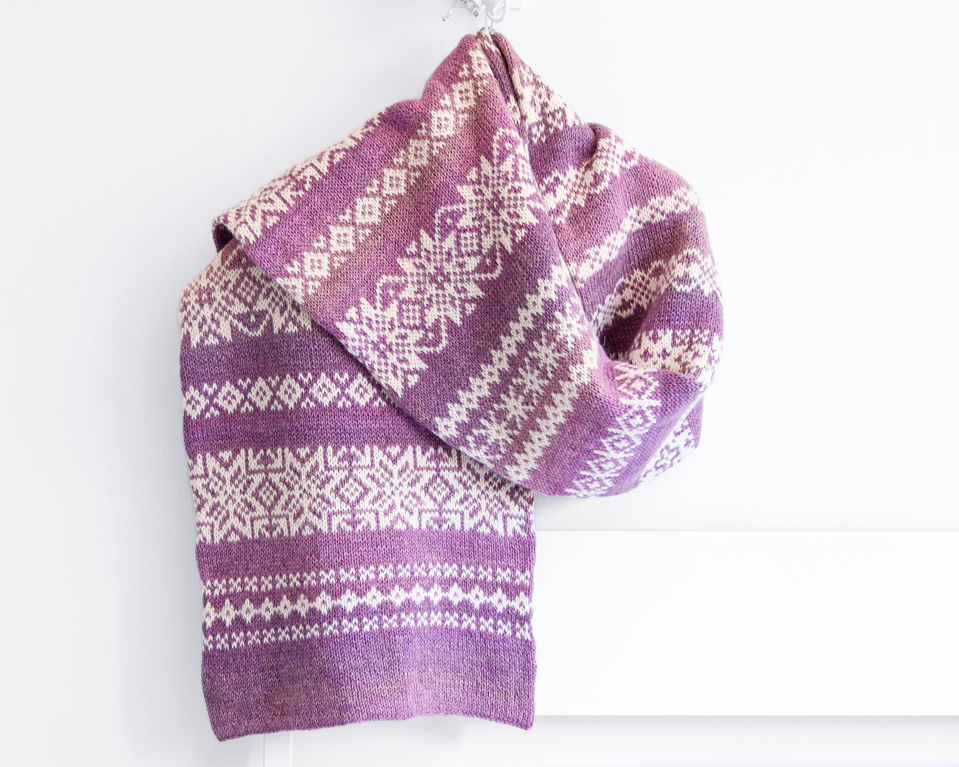 A purple and white hand-knitted Fair Isle scarf draped over a white shelf against a white wall.