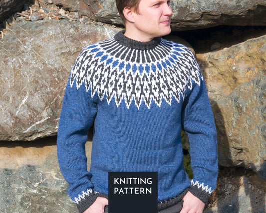 A man wearing a blue, white, and gray Odin icelandic lopapeysa sweater with a classic yoke, standing outdoors in front of large rocks.