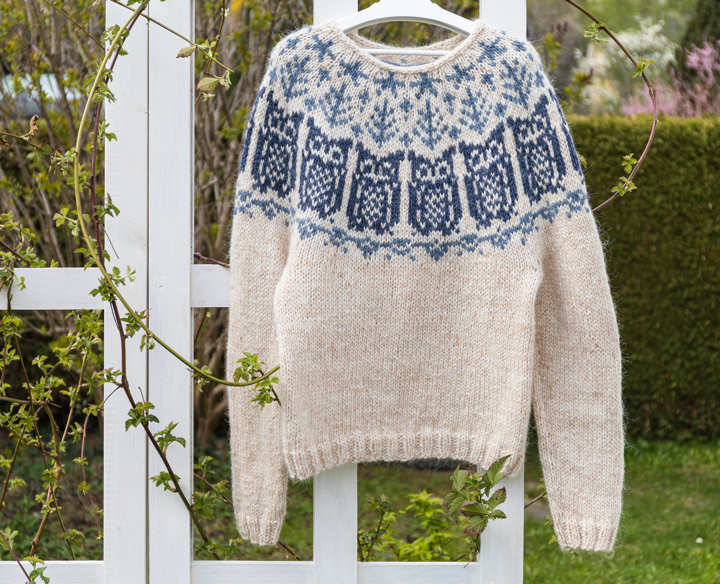 A hand-knitted wool lopapeysa sweater featuring an owl pattern on the yoke, displayed on a white frame with foliage in the background.