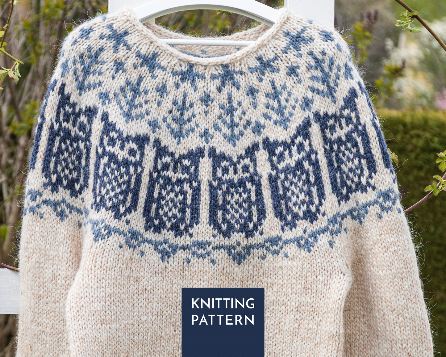 A cream-colored knit sweater with a blue and navy owls yoke pattern, hanging on a white hanger outdoors.