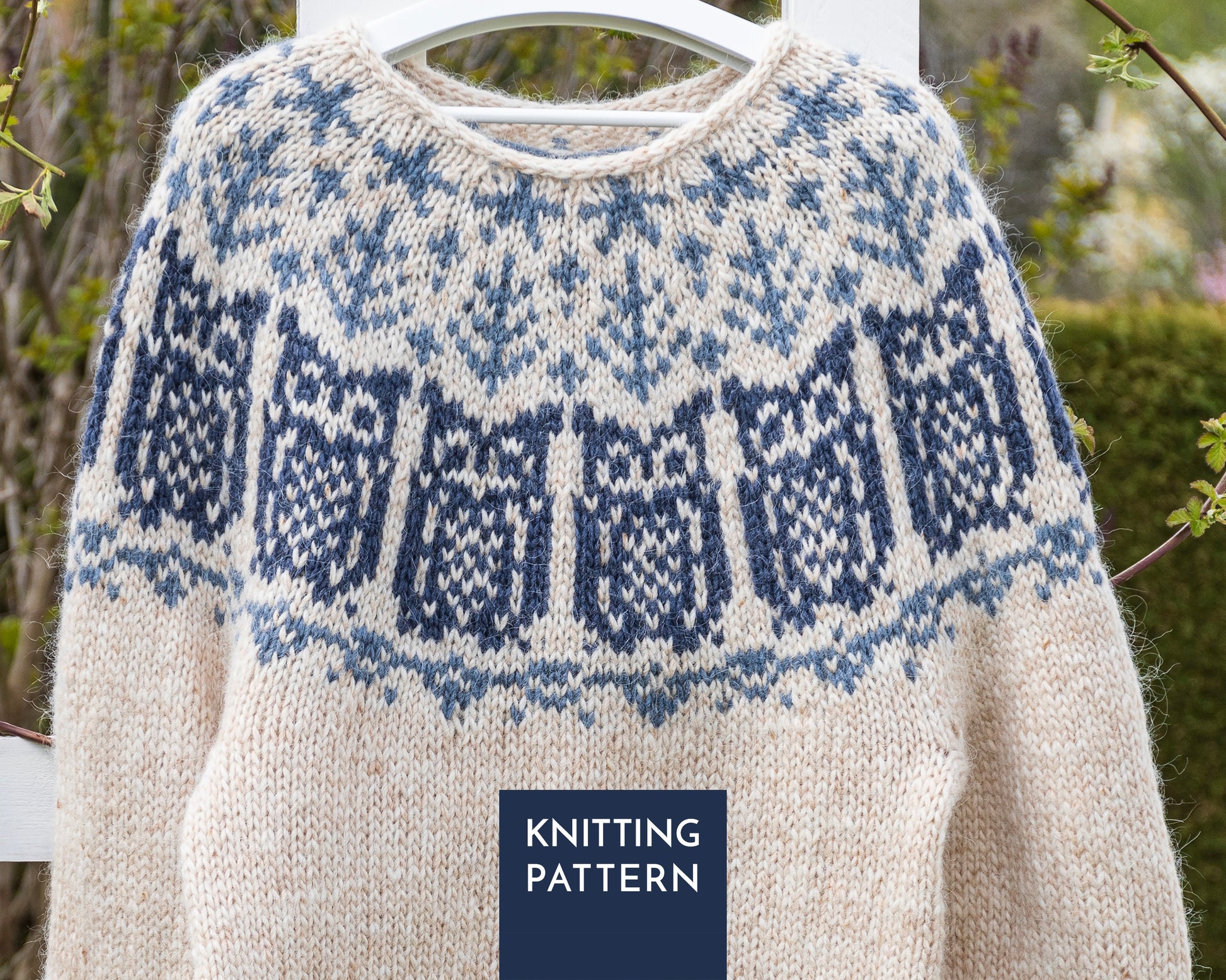 A cream-colored knit sweater with a blue and navy owls yoke pattern, hanging on a white hanger outdoors.