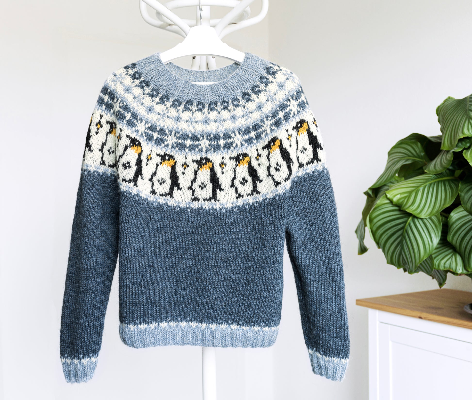 A hand-knitted Icelandic lopapeysa sweater featuring a penguin pattern, displayed against a white background with a green plant to the side.
