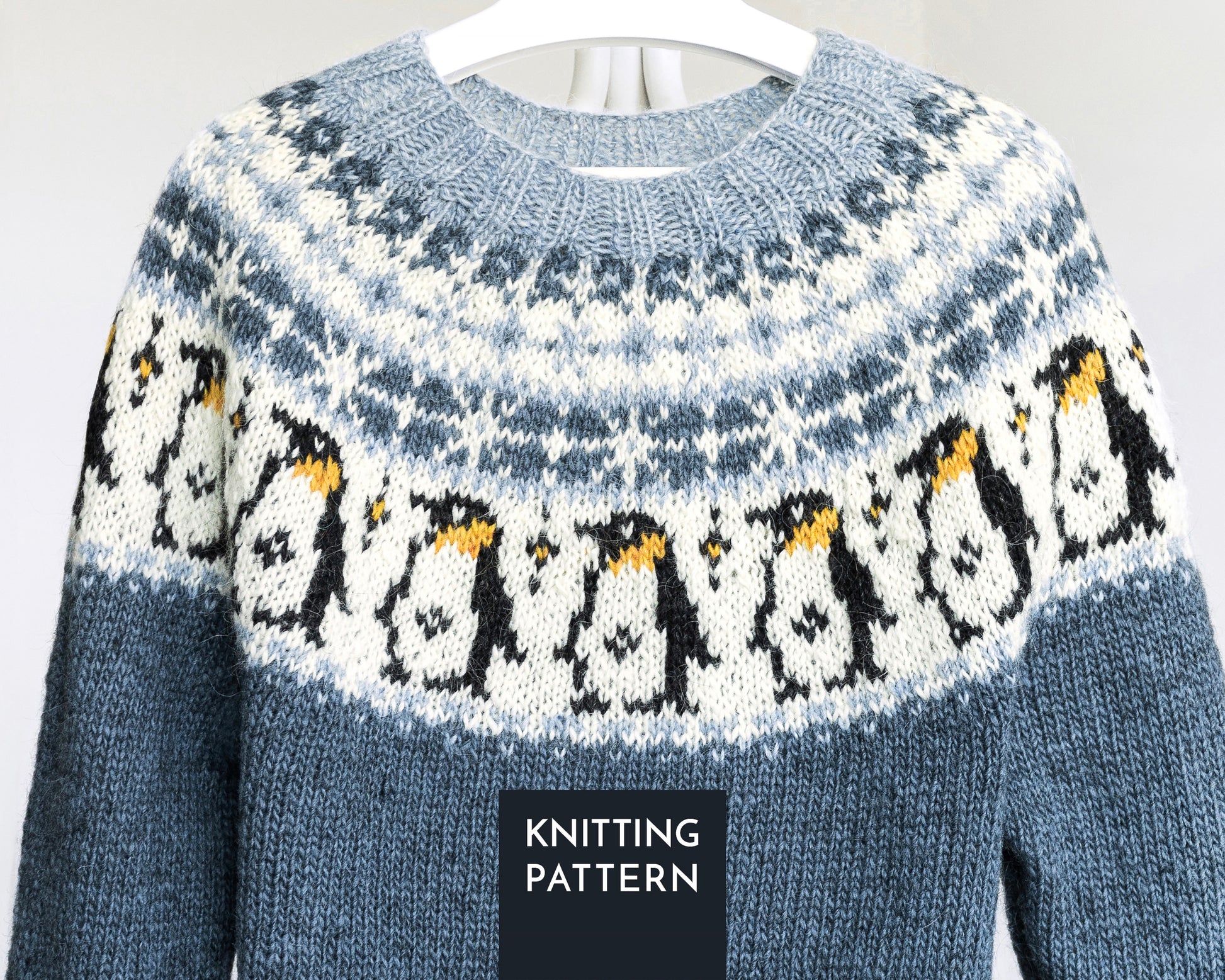 A knitted sweater shown on a hanger against a plain light background. The sweater has a blue-gray base with a white yoke featuring a penguin fair isle pattern in black, white, and yellow.