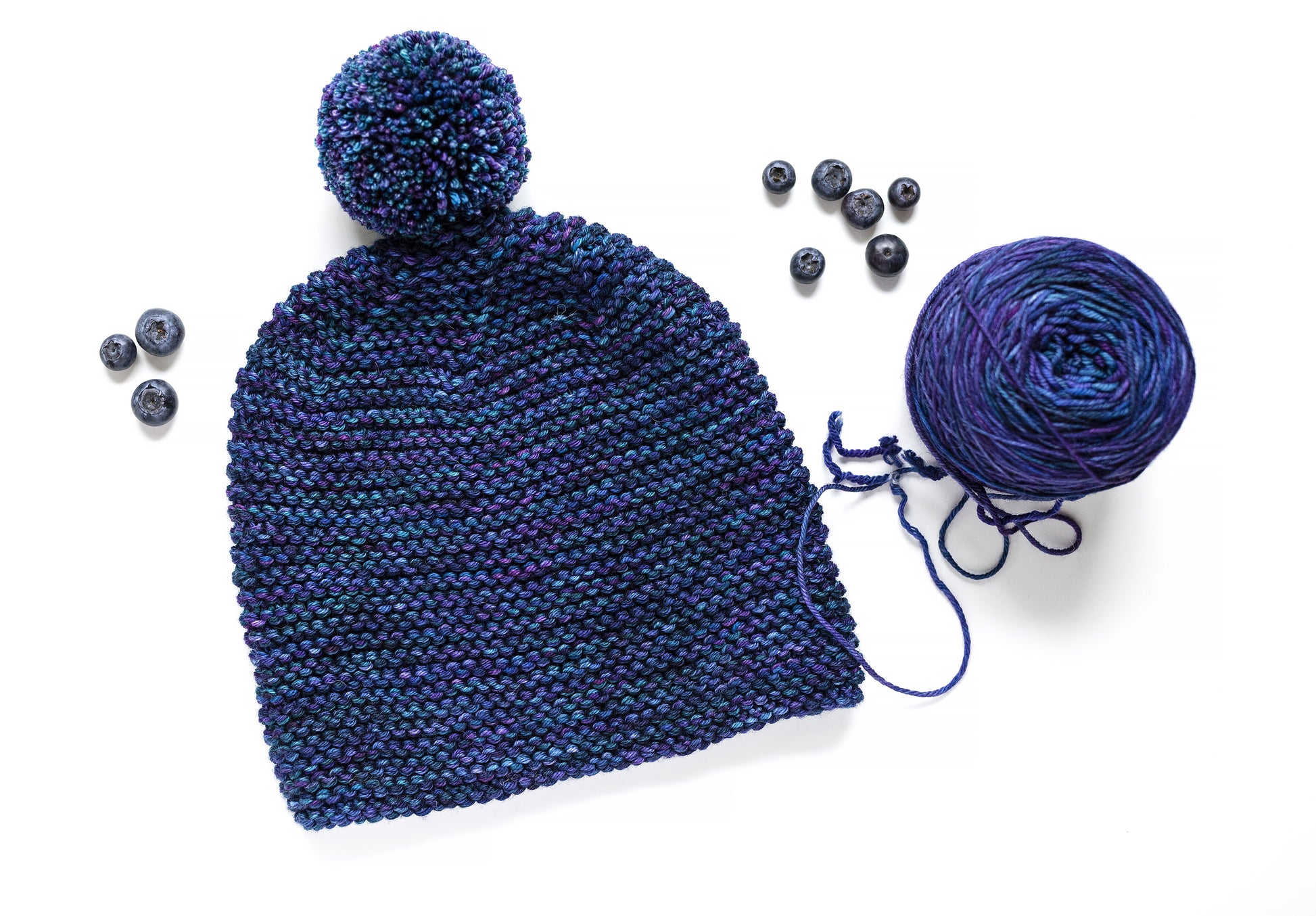 Front view of a blue and purple gradient wool garter stitch hand-knitted pom pom hat with yarn ball