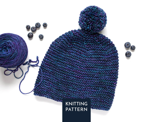 A knitted pom-pom beanie in a variegated navy and purple yarn, styled with a ball of matching yarn and blueberries on a white background, with a 'KNITTING PATTERN' label overlay.