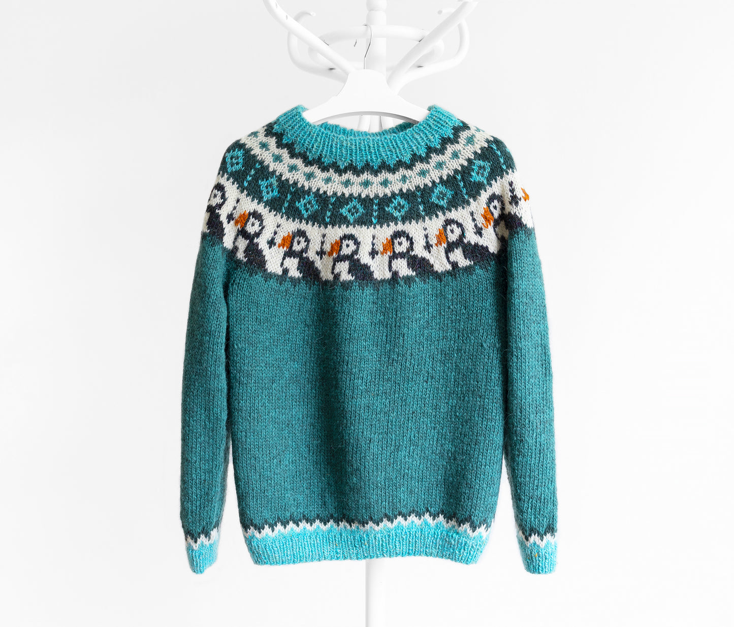 Front view of a teal Icelandic lopapeysa sweater with a colorful puffin yoke pattern, hanging on a white hanger against a white background.
