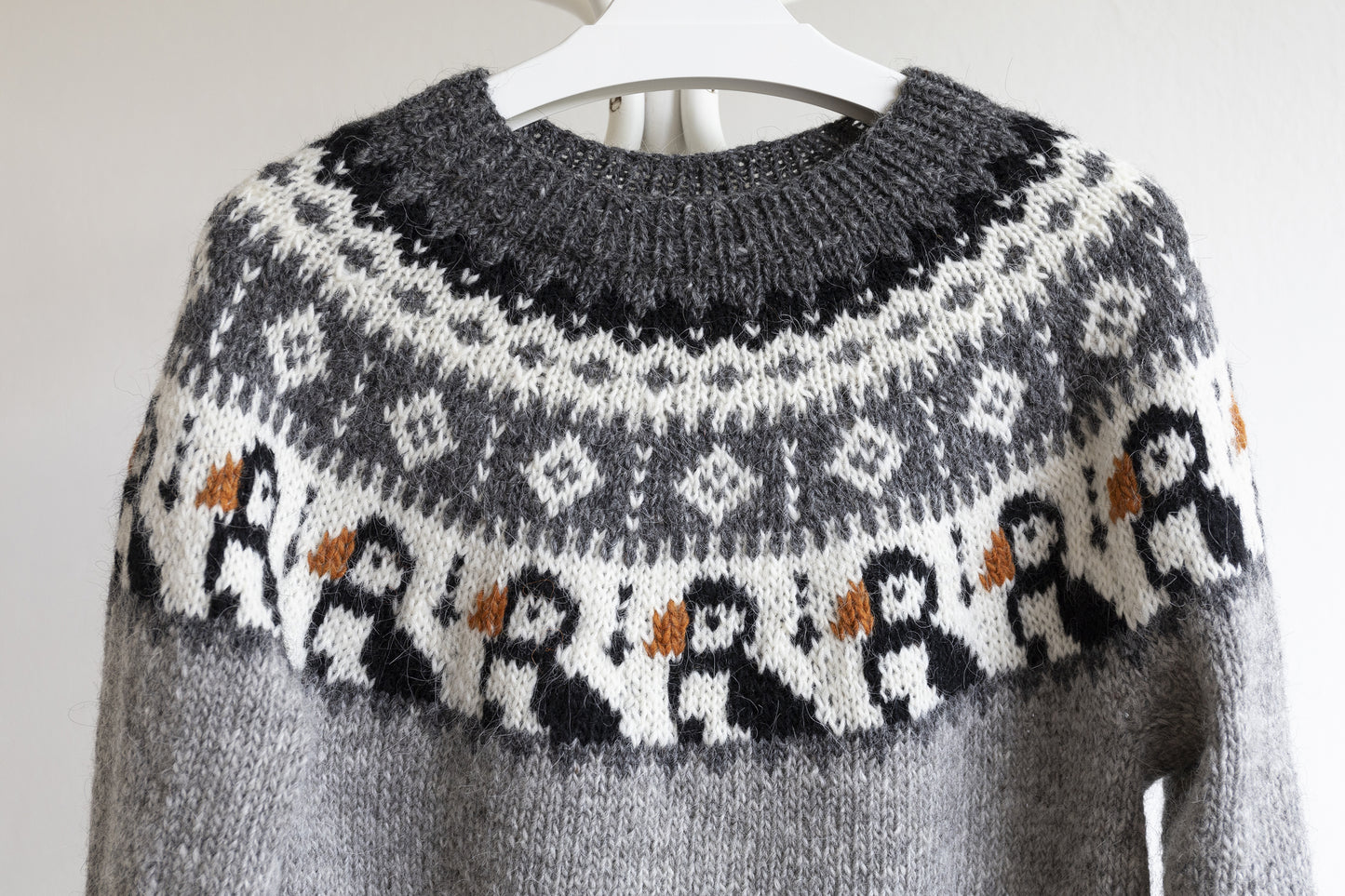 A gray knit sweater with a white Fair Isle yoke featuring puffin motifs with orange beaks, hanging on a white hanger against a light background.