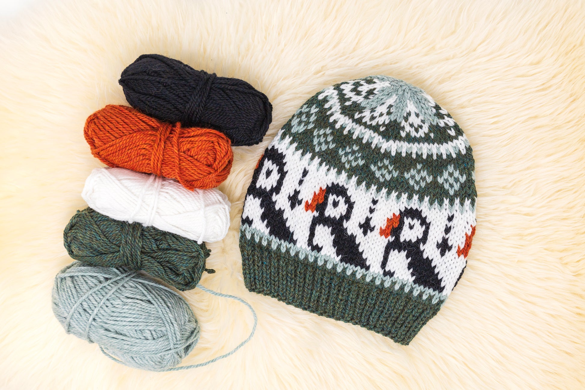 a hand-knitted wool hat with a geometric pattern featuring puffins in green, black and white colors on a white background accompanied by yarn skeins