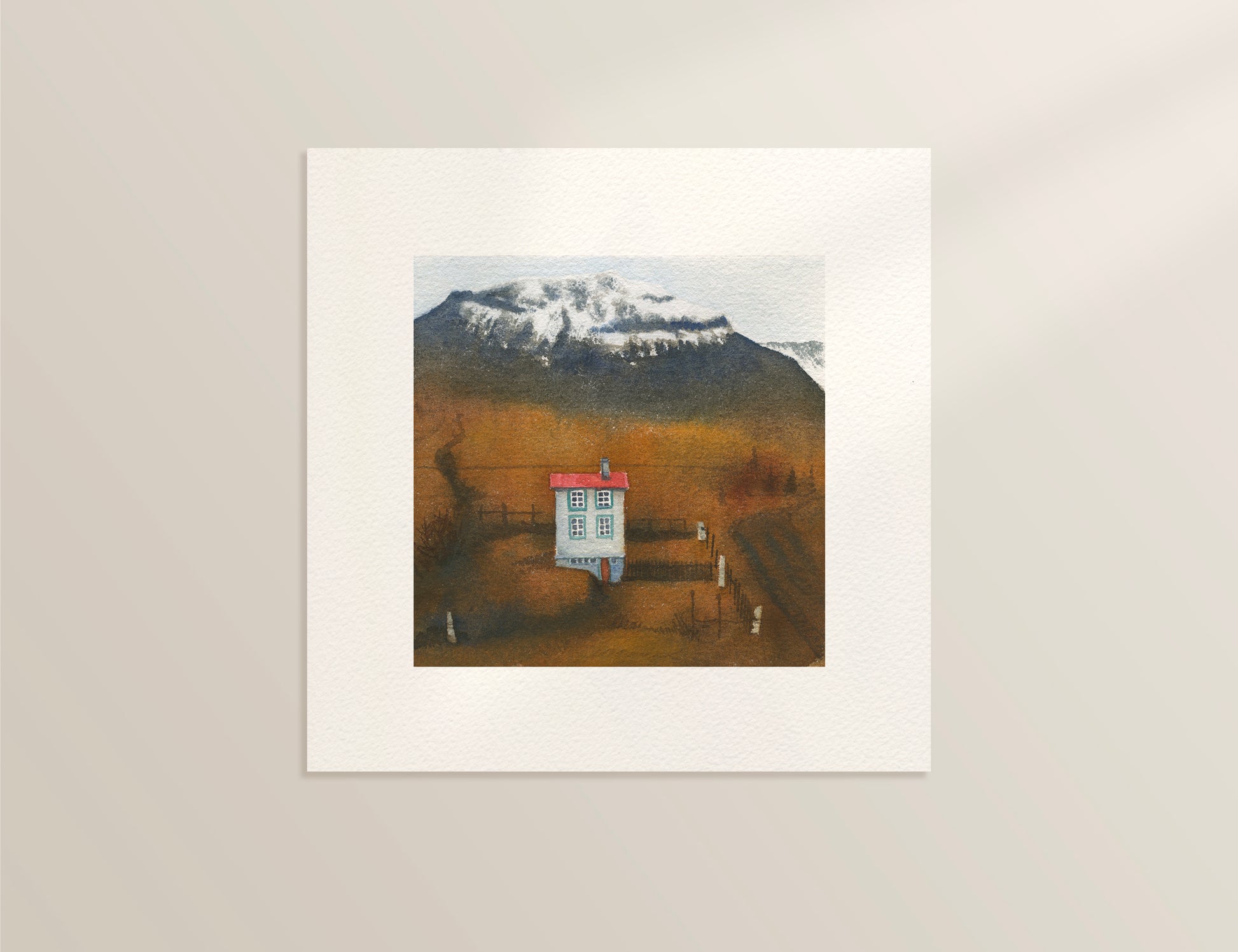Framed artwork of a house with mountains in the background on a beige wall.