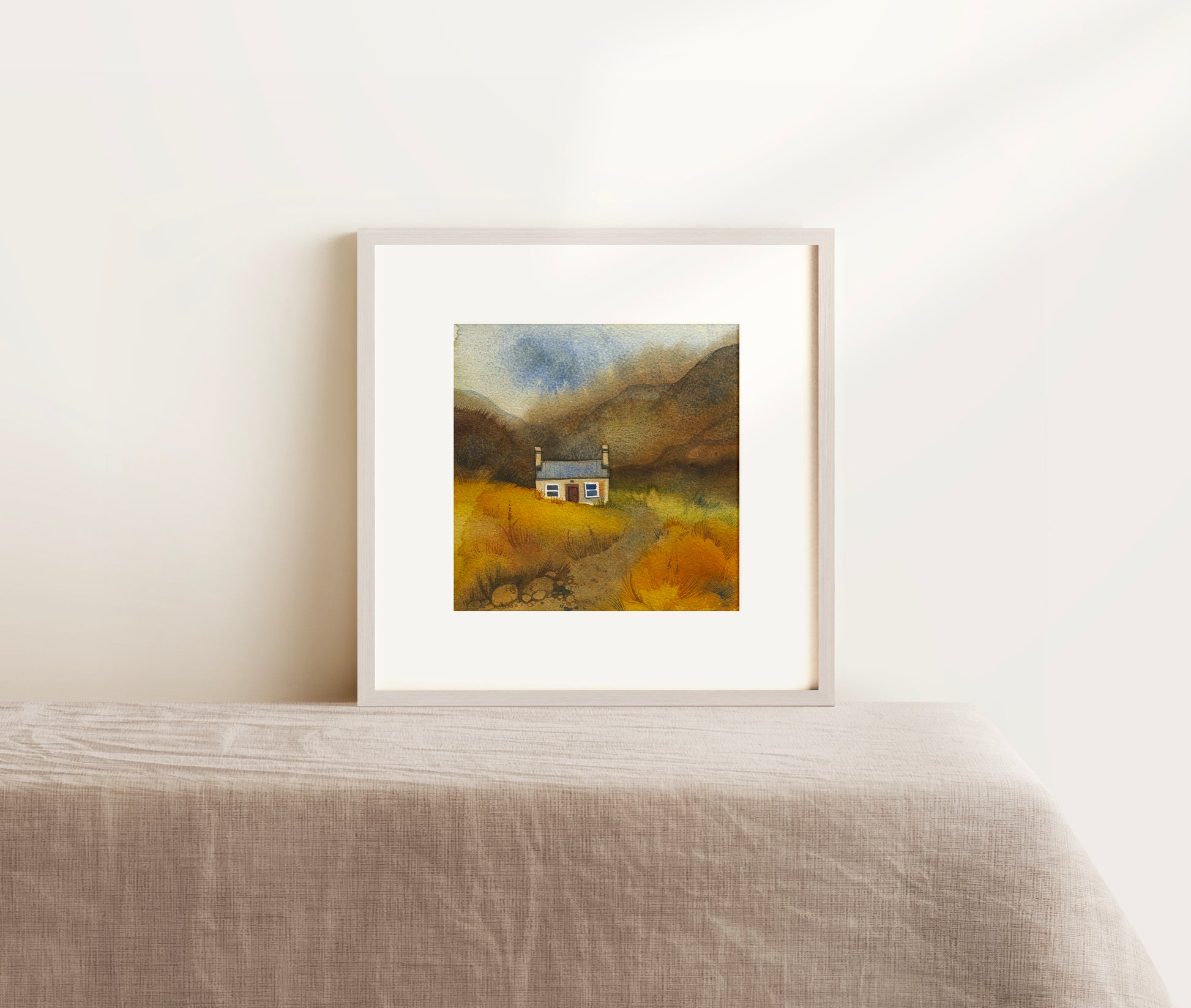 Framed watercolor landscape painting of a house in a field on a white wall.