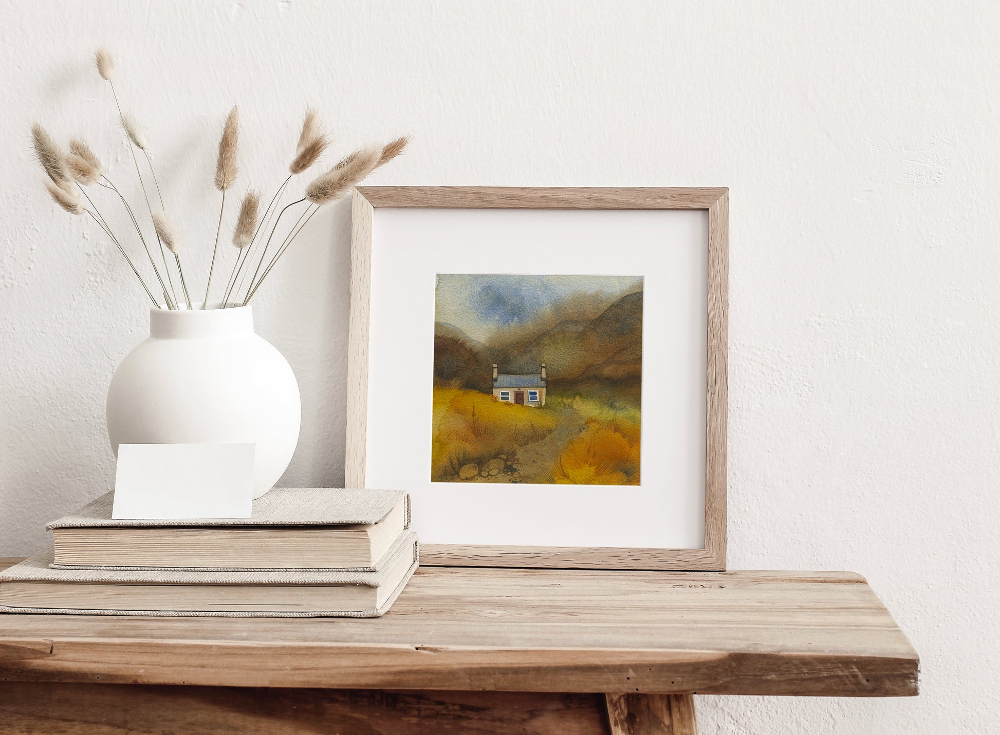 Framed watercolor landscape painting on a wooden surface with a white vase and books.