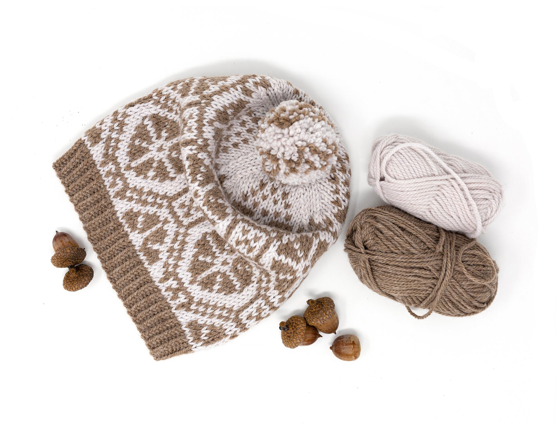 Hand-knitted pom-pom hat with carriage wheel pattern, yarn balls, and acorns on a white background