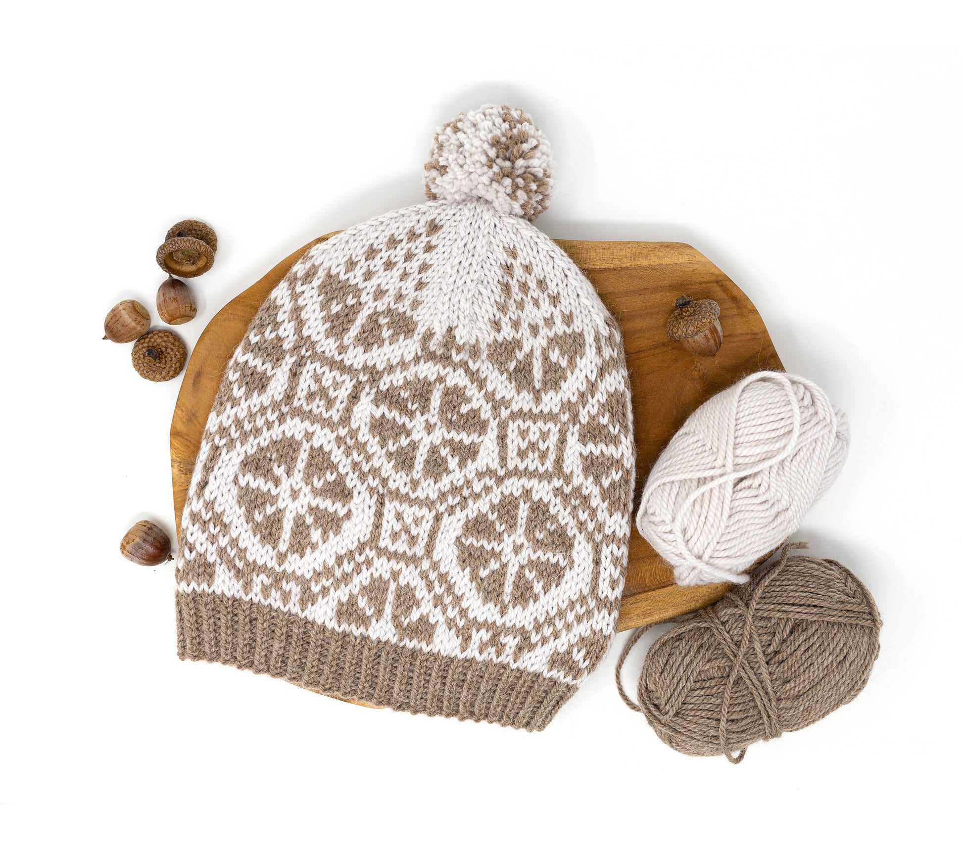 Hand-knitted Ratas Fair Isle pom pom hat with a pattern, yarn balls, and acorns on a wooden board.