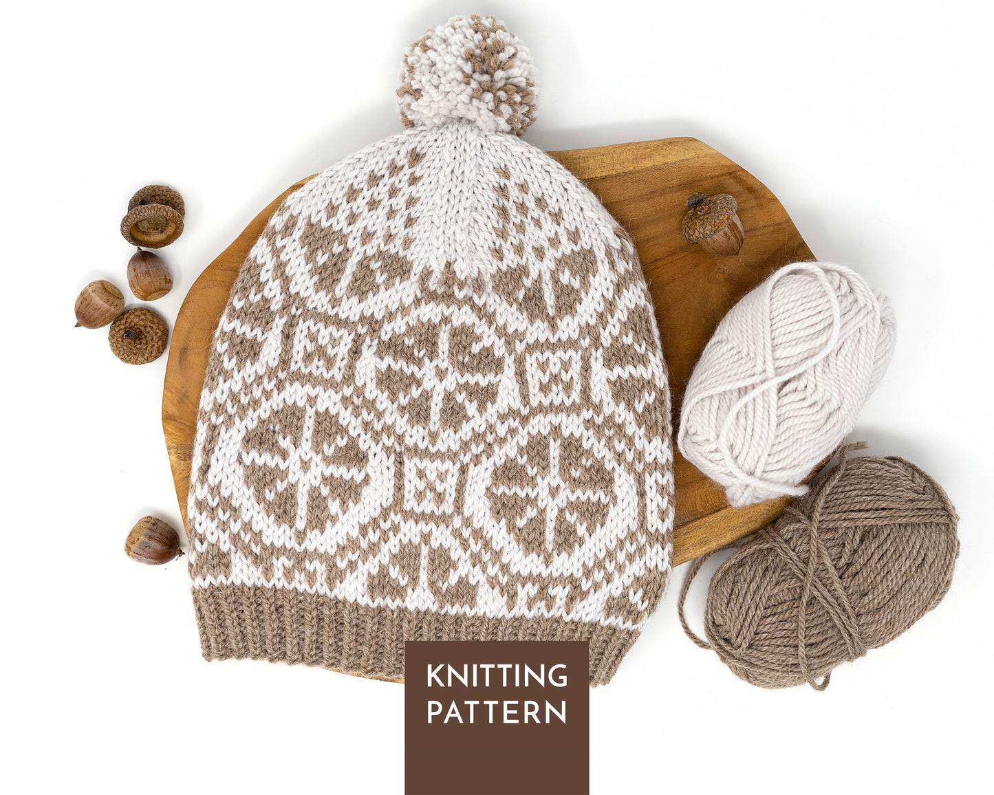 A knitted pom-pom beanie with a brown and off-white Fair Isle wheels pattern, styled on a wooden board with yarn balls and acorns, with a 'KNITTING PATTERN' label overlay. 