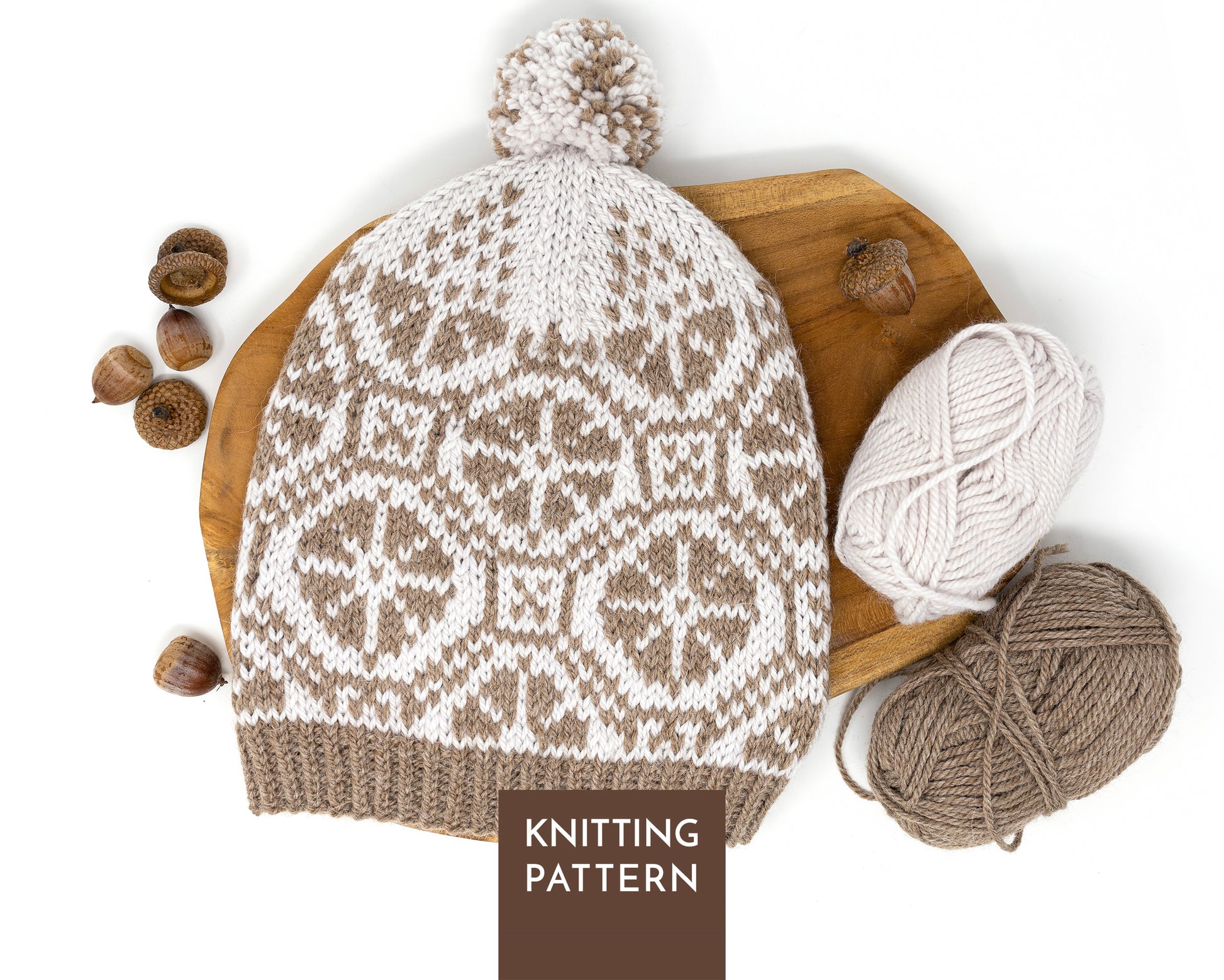 A knitted pom-pom beanie with a brown and off-white Fair Isle wheels pattern, styled on a wooden board with yarn balls and acorns, with a 'KNITTING PATTERN' label overlay. 