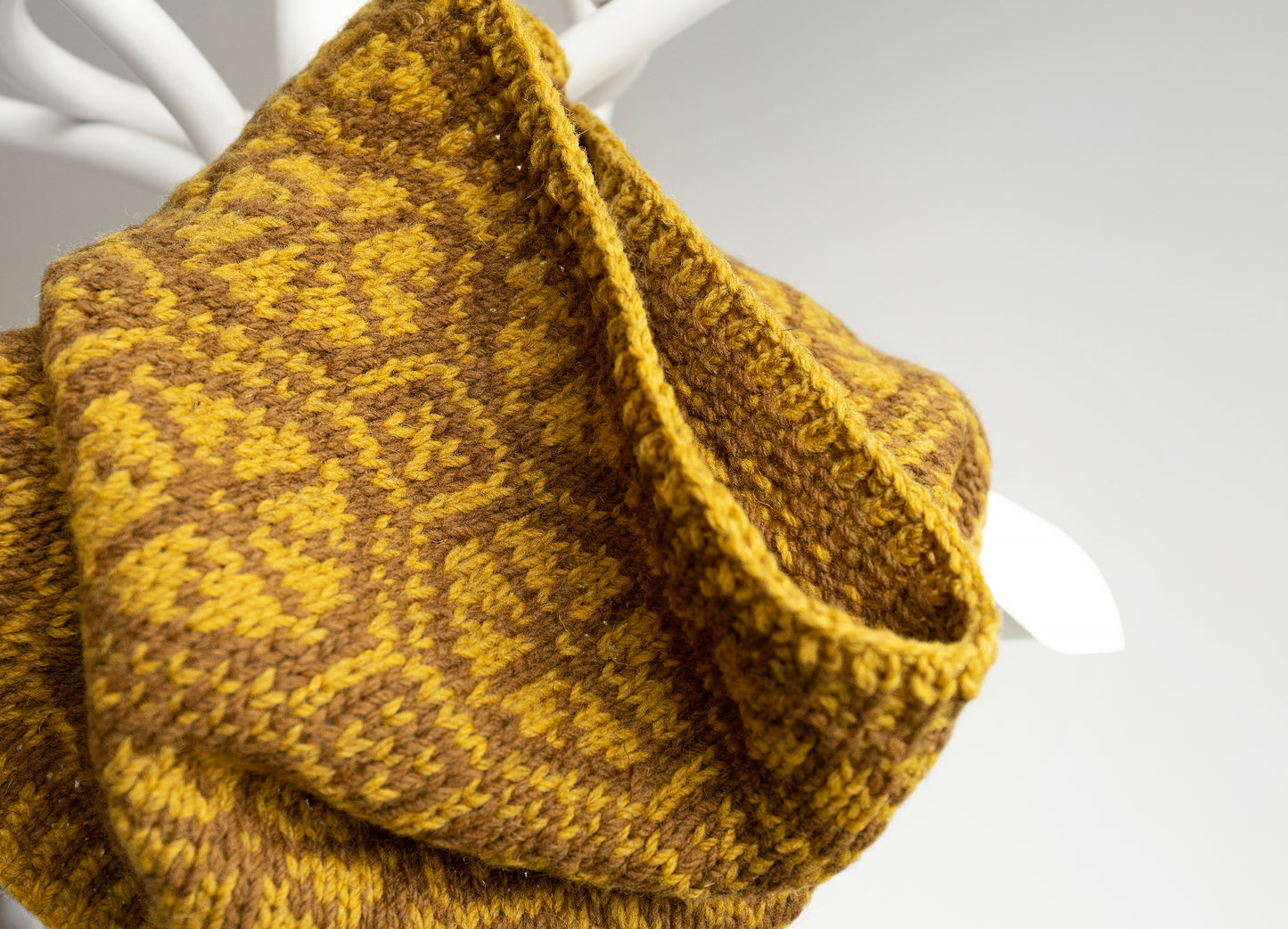 Hand-knitted mustard yellow cowl on a white stand against a light gray background
