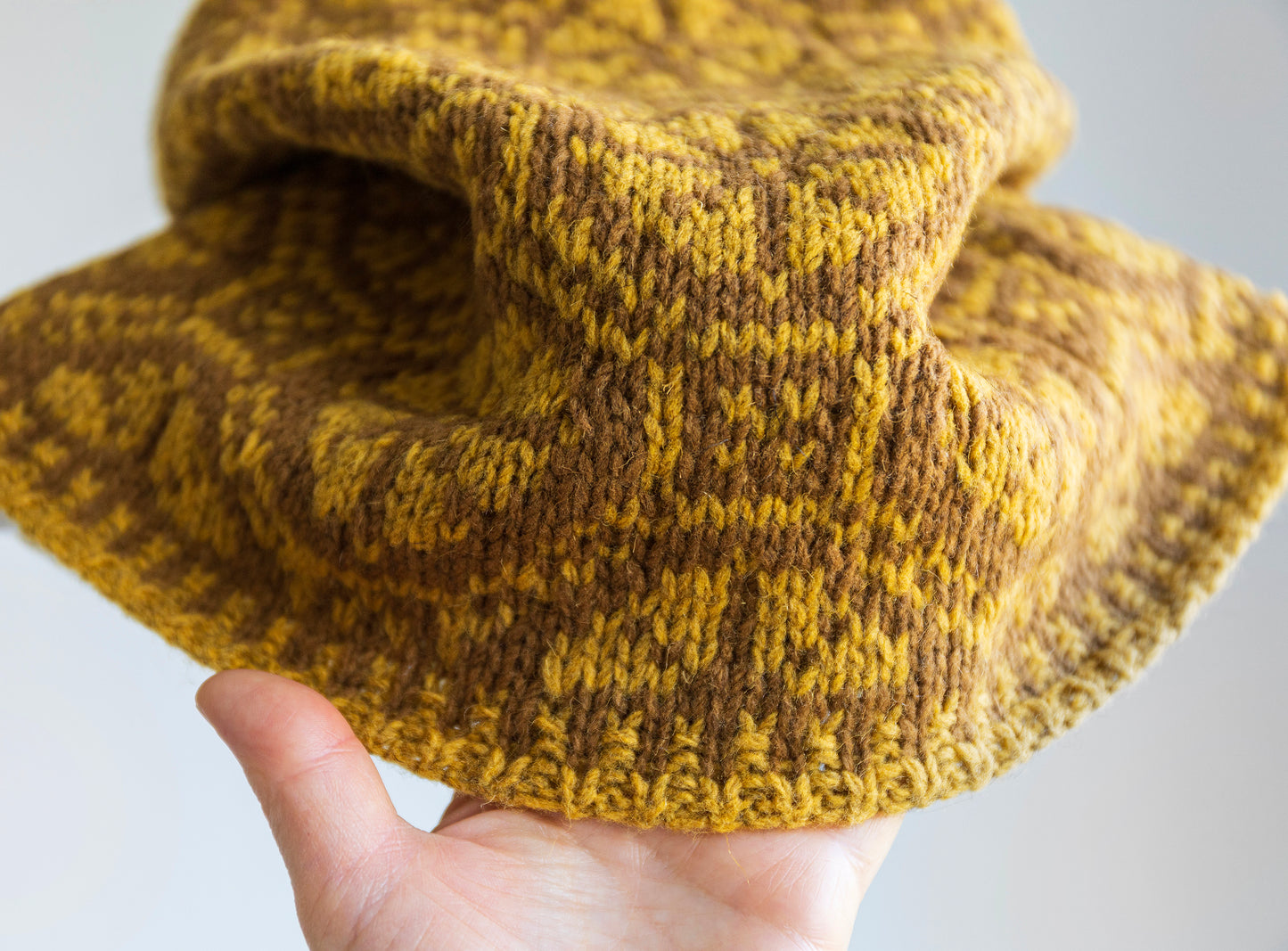 Hand holding a knitted yellow and brown Ratas cowl scarf against a light background