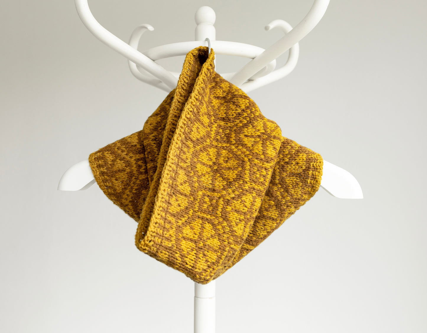 Mustard yellow hand-knitted Ratas  cowl scarf on a white coat rack against a light gray background