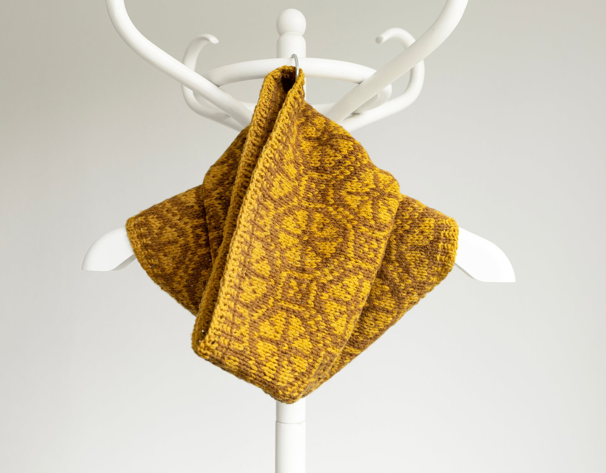 Mustard yellow hand-knitted Ratas  cowl scarf on a white coat rack against a light gray background