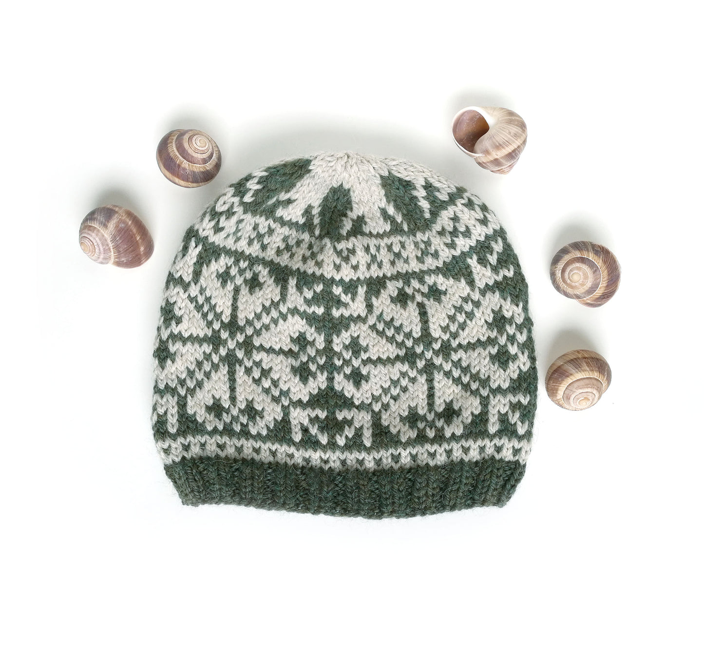 A dark green and cream knit beanie hat with a traditional Nordic snowflake pattern, styled flat-lay on a white background with five small seashells arranged around it.