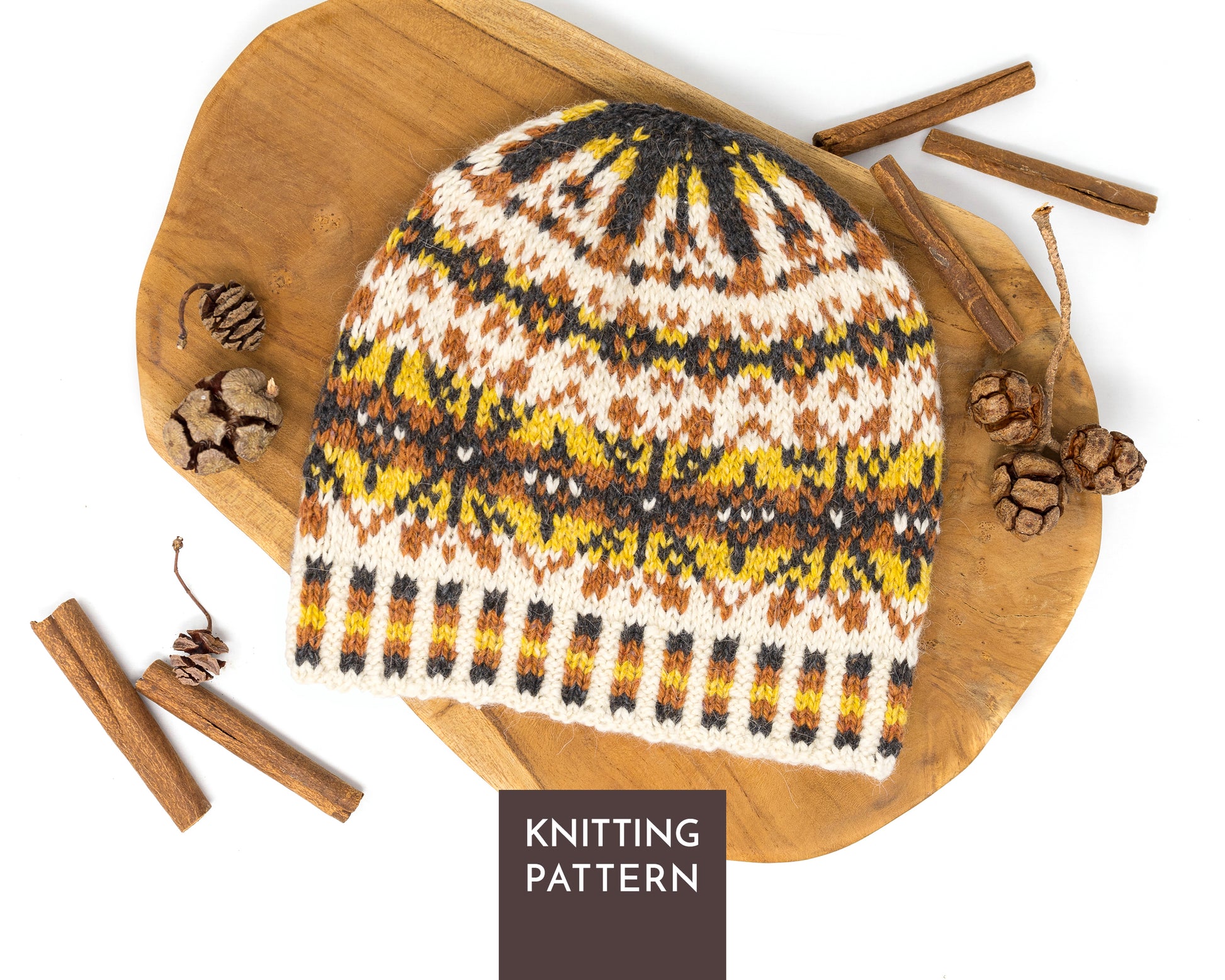 A knitted beanie hat with a Fair Isle pattern in cream, yellow, brown, and black, displayed on a wooden board with cinnamon sticks and pinecones. A dark brown label at the bottom reads 'KNITTING PATTERN'.