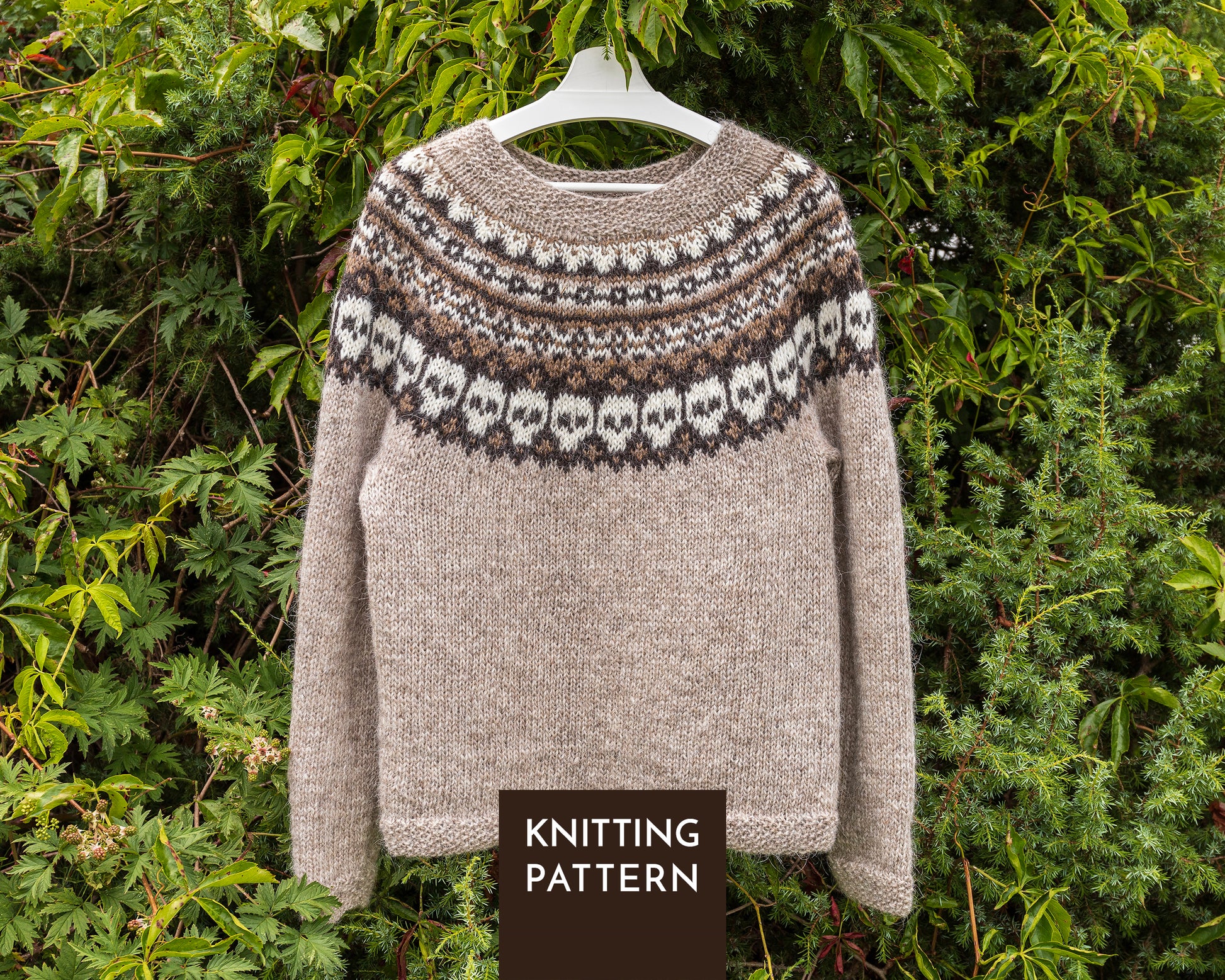 A knitted sweater with a beige body and a brown and white Fair Isle skull yoke pattern, hanging on a white hanger against a background of green foliage. A dark brown label at the bottom reads 'KNITTING PATTERN'.