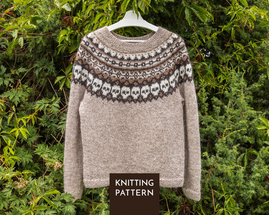 A knitted sweater with a beige body and a brown and white Fair Isle skull yoke pattern, hanging on a white hanger against a background of green foliage. A dark brown label at the bottom reads 'KNITTING PATTERN'.