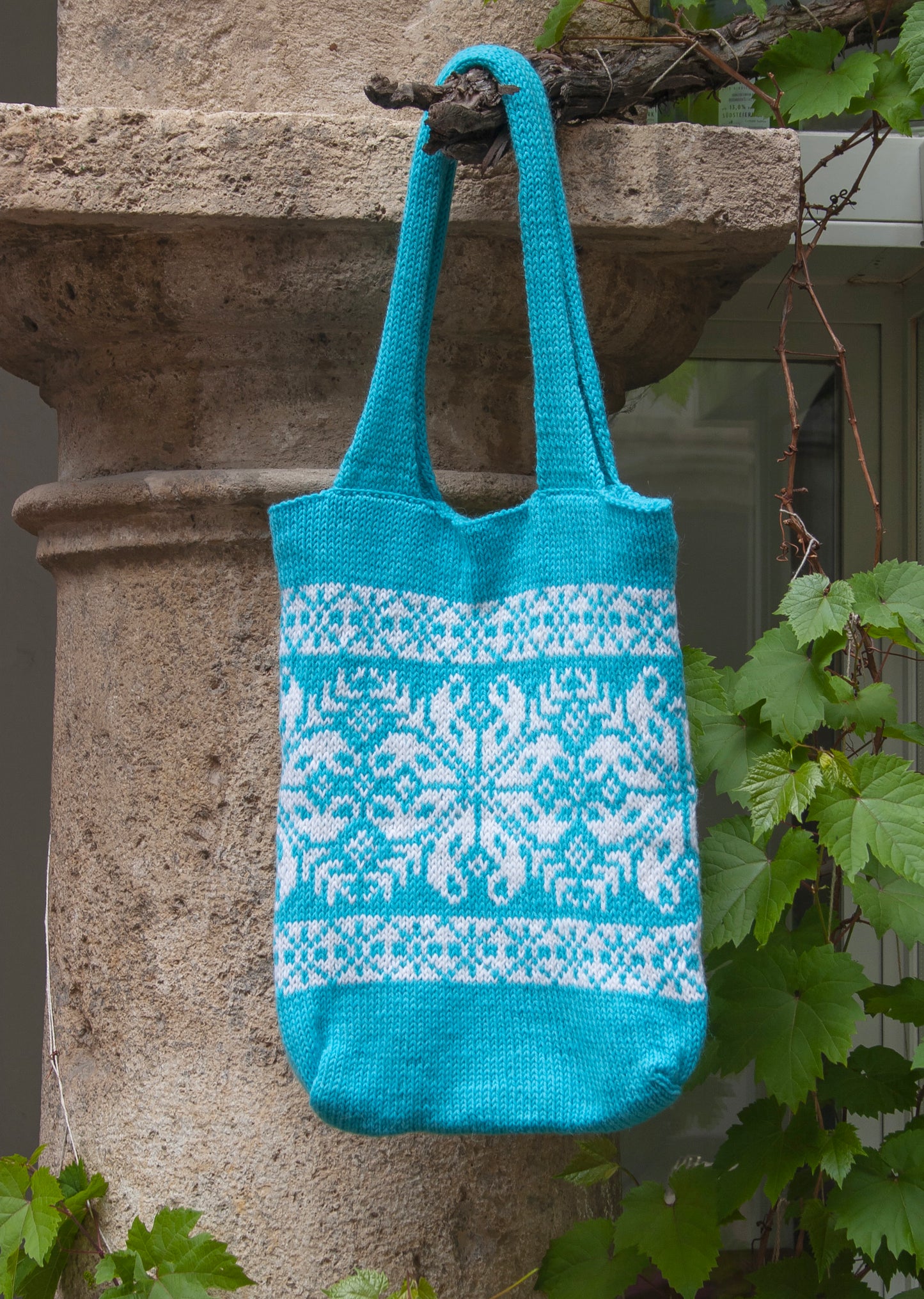 Turquoise and white Snowflake Selbu hand-knitted tote bag hanging on a grapevine
