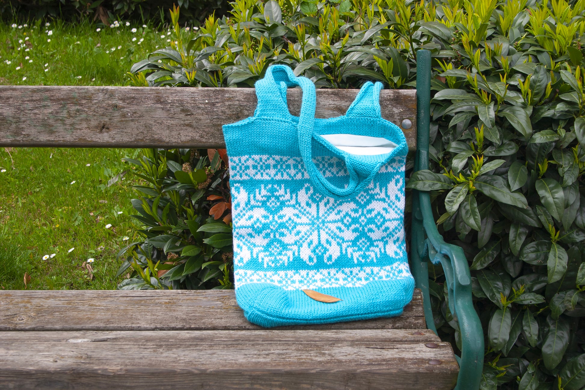 A turquoise hand-knitted tote bag with a snowflake pattern, placed on a wooden bench with green foliage in the background.