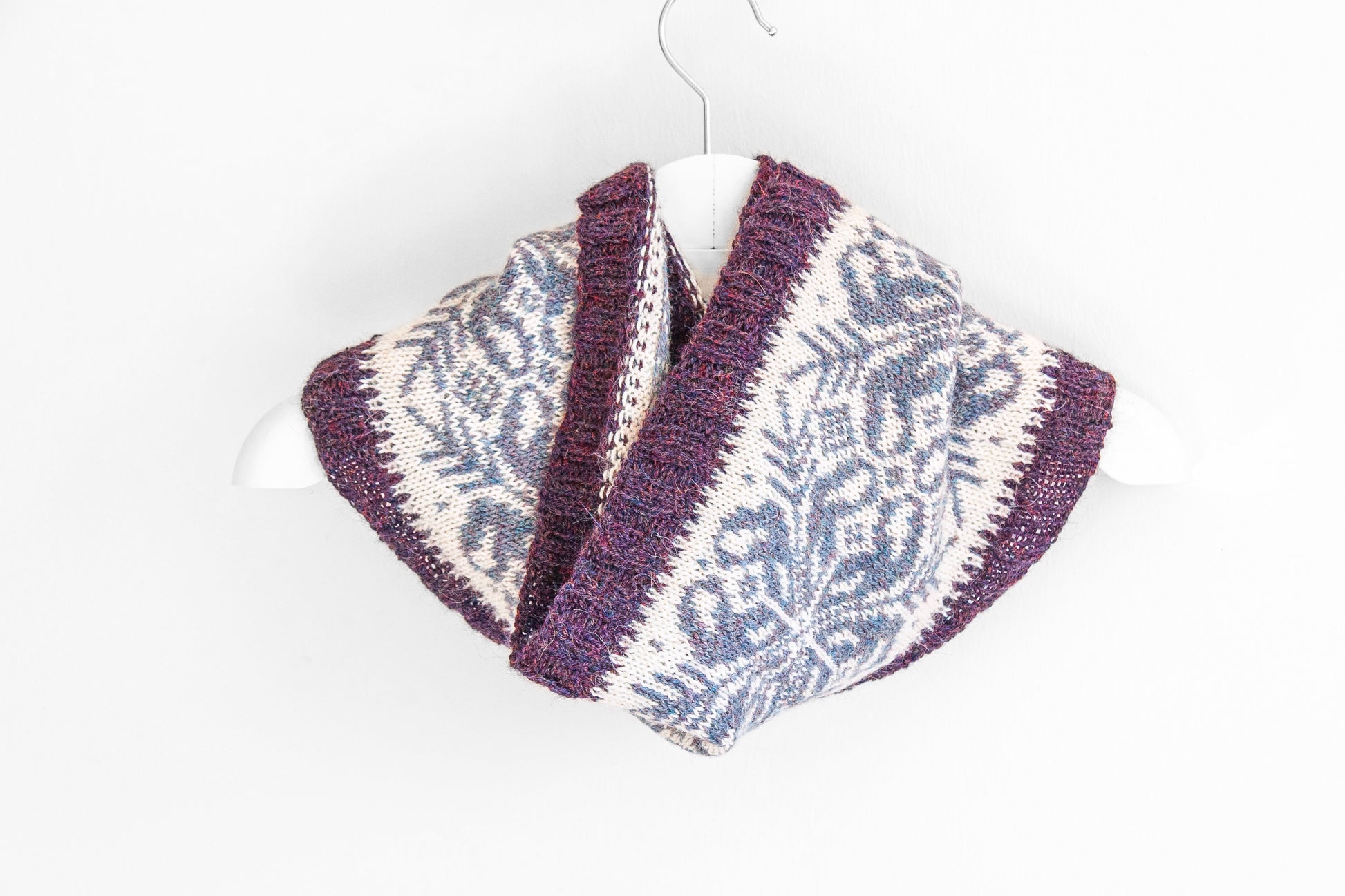 A knitted cowl scarf with a purple and cream Fair Isle snowflake pattern, draped on a white hanger against a plain white background.