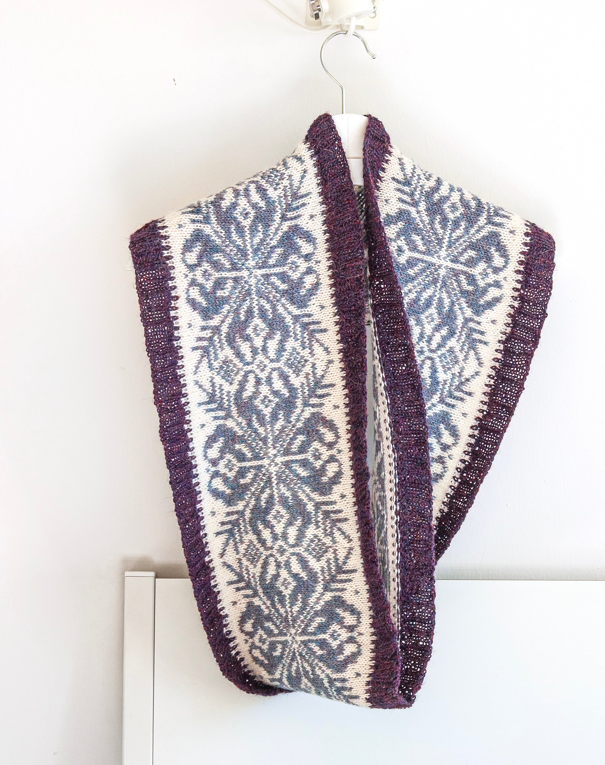 A knitted infinity scarf with a dark purple border and a central cream and slate blue-gray snowflake pattern, hanging on a white hanger against a white wall.