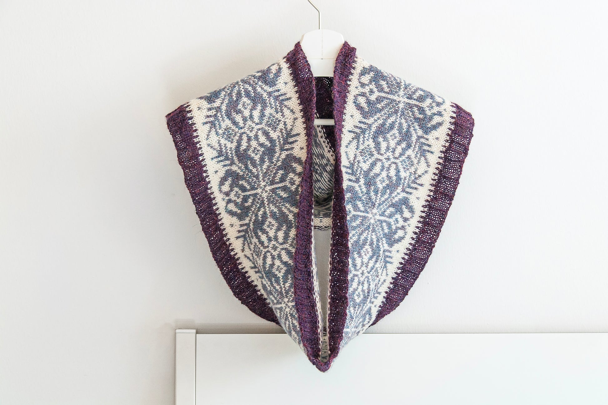Hand-knitted alpaca infinity scarf with a purple, gray and cream Fair Isle snowflake pattern, hanging on a white hanger against a plain white wall.
