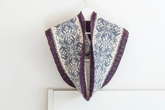 Hand-knitted alpaca infinity scarf with a purple, gray and cream Fair Isle snowflake pattern, hanging on a white hanger against a plain white wall.