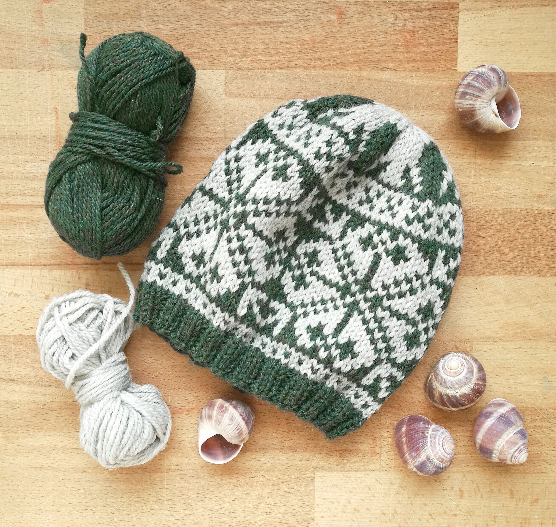 Knitted hat with green and white pattern on a wooden surface with yarn and shells.
