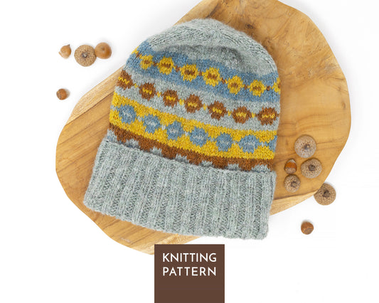 A knitted beanie hat with a gray folded brim and a multicolor Fair Isle pattern in blue, yellow, and brown, displayed on a wooden board with acorns. A brown label at the bottom reads 'KNITTING PATTERN'.
