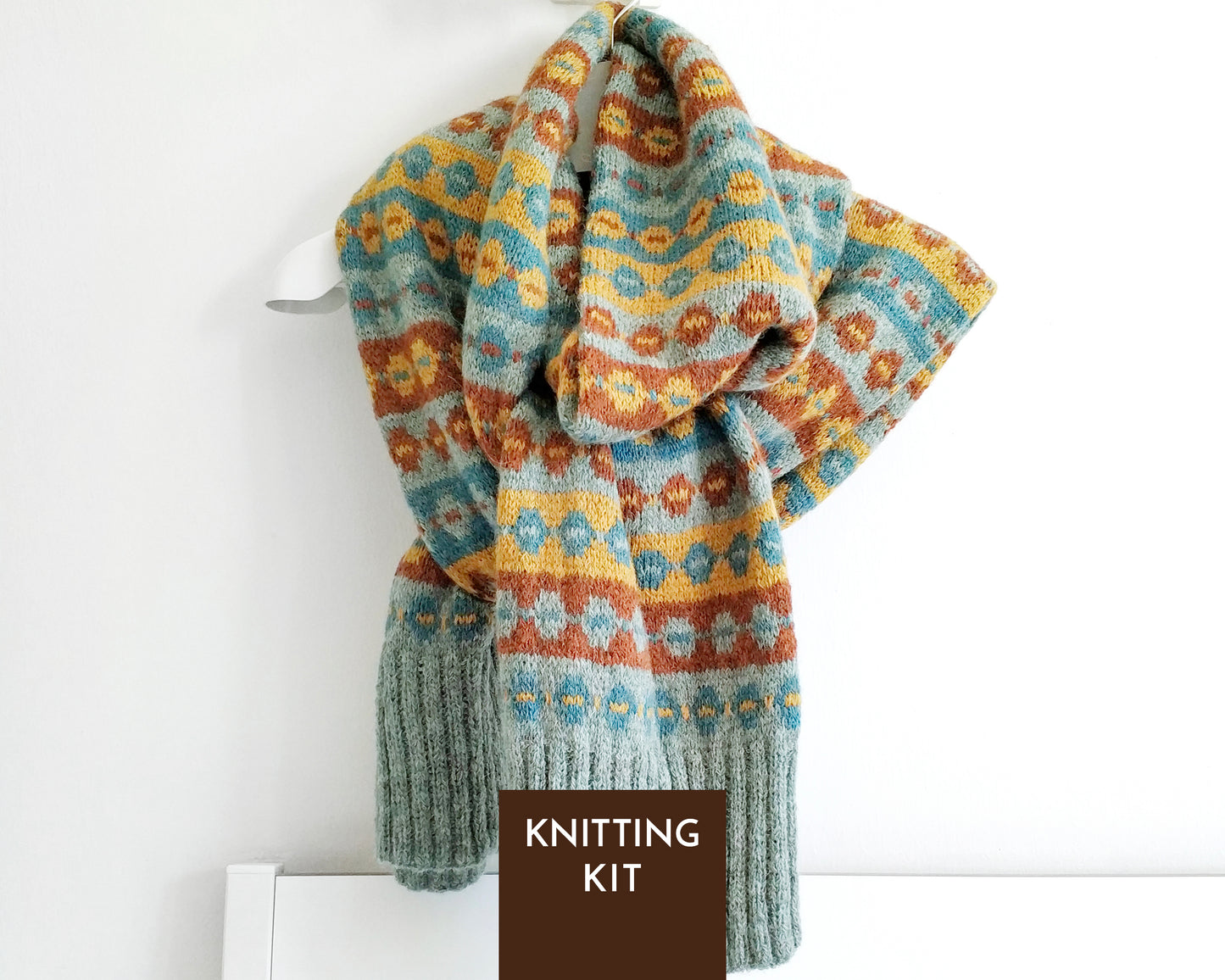 A knitted scarf with a multicolor Fair Isle pattern in blue, yellow, and brown, draped on a white hanger against a white wall. A dark brown label at the bottom reads 'KNITTING KIT'.