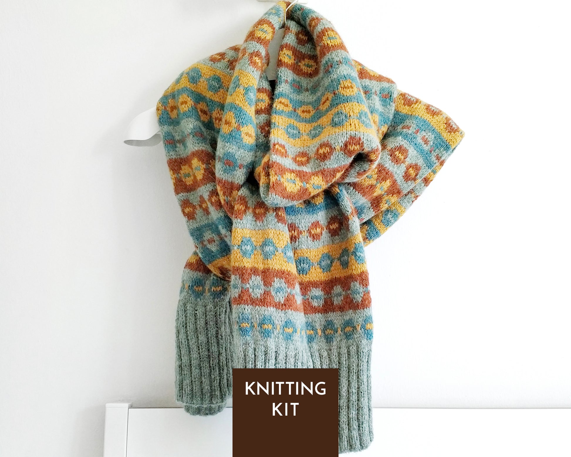 A knitted scarf with a multicolor Fair Isle pattern in blue, yellow, and brown, draped on a white hanger against a white wall. A dark brown label at the bottom reads 'KNITTING KIT'.