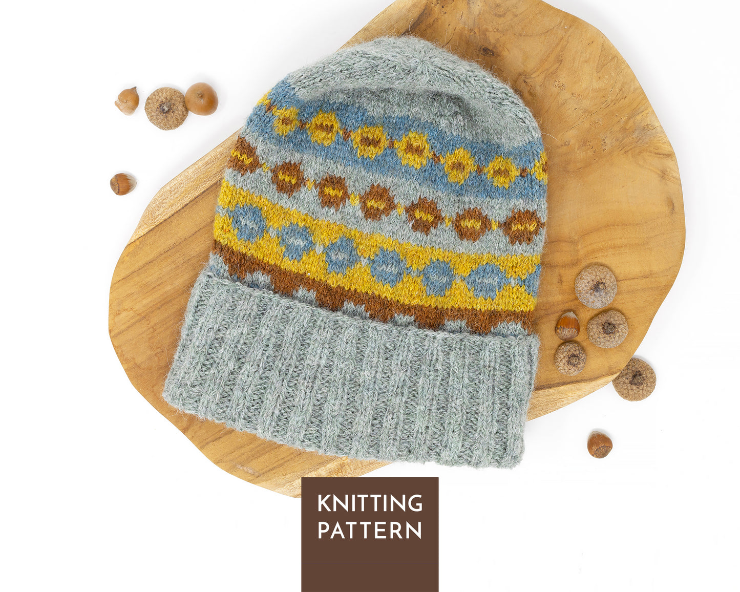 A knitted beanie hat with a gray ribbed cuff and a multicolor Fair Isle pattern in blue, yellow, and brown, displayed on a wooden board with acorns. A brown label at the bottom reads 'KNITTING PATTERN'.