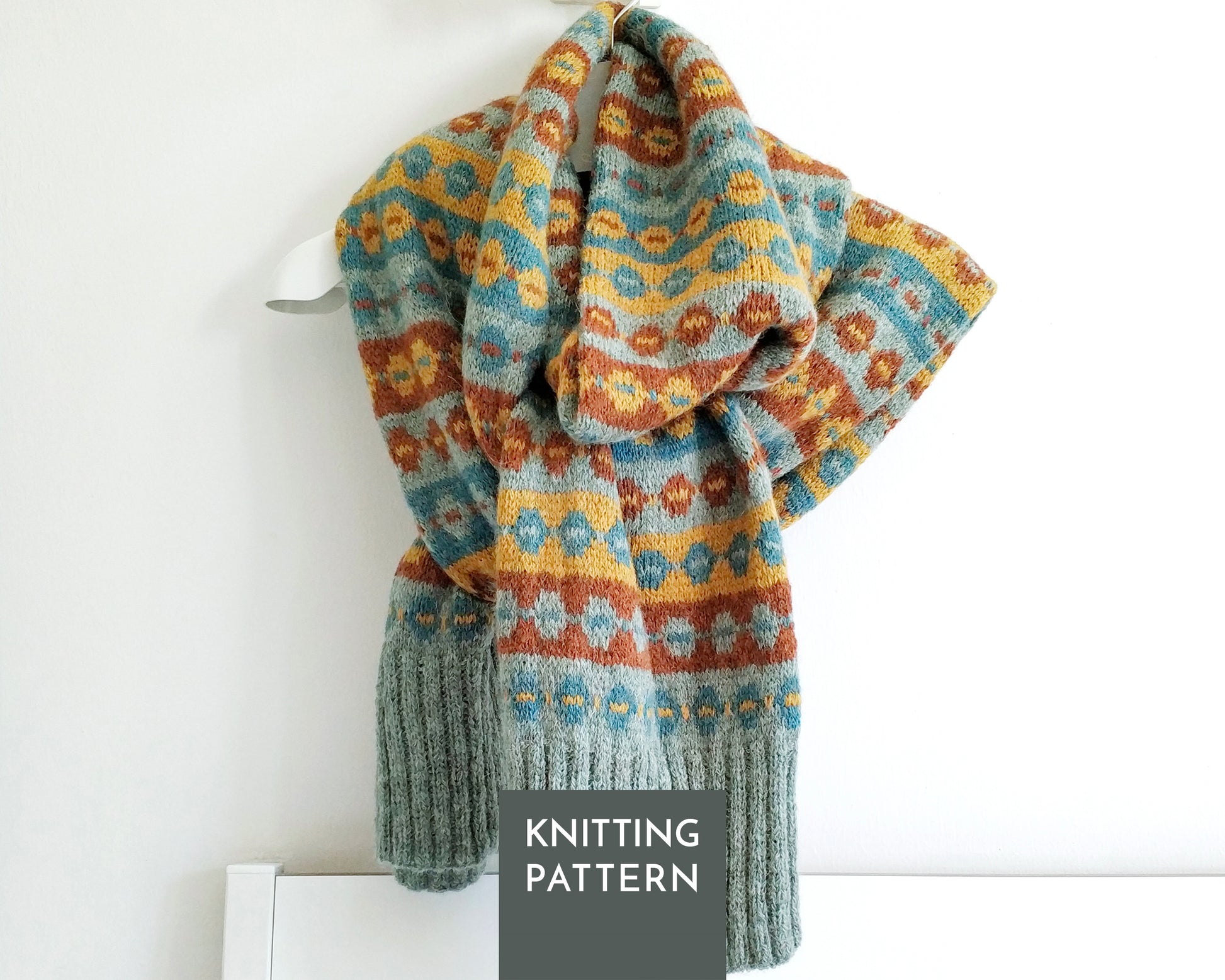 A knitted scarf with a multicolor Fair Isle pattern in blue, yellow, and brown, draped over a white hanger against a white wall. A dark gray label at the bottom reads 'KNITTING PATTERN'.