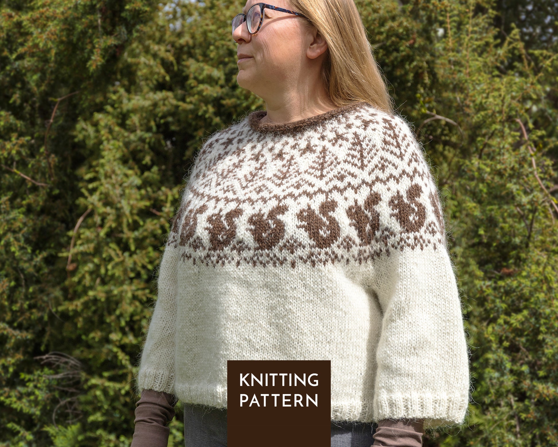 A woman wearing a knitted sweater with a cream body and brown colorwork yoke with squirrels and trees across the chest, standing outdoors in front of green trees.