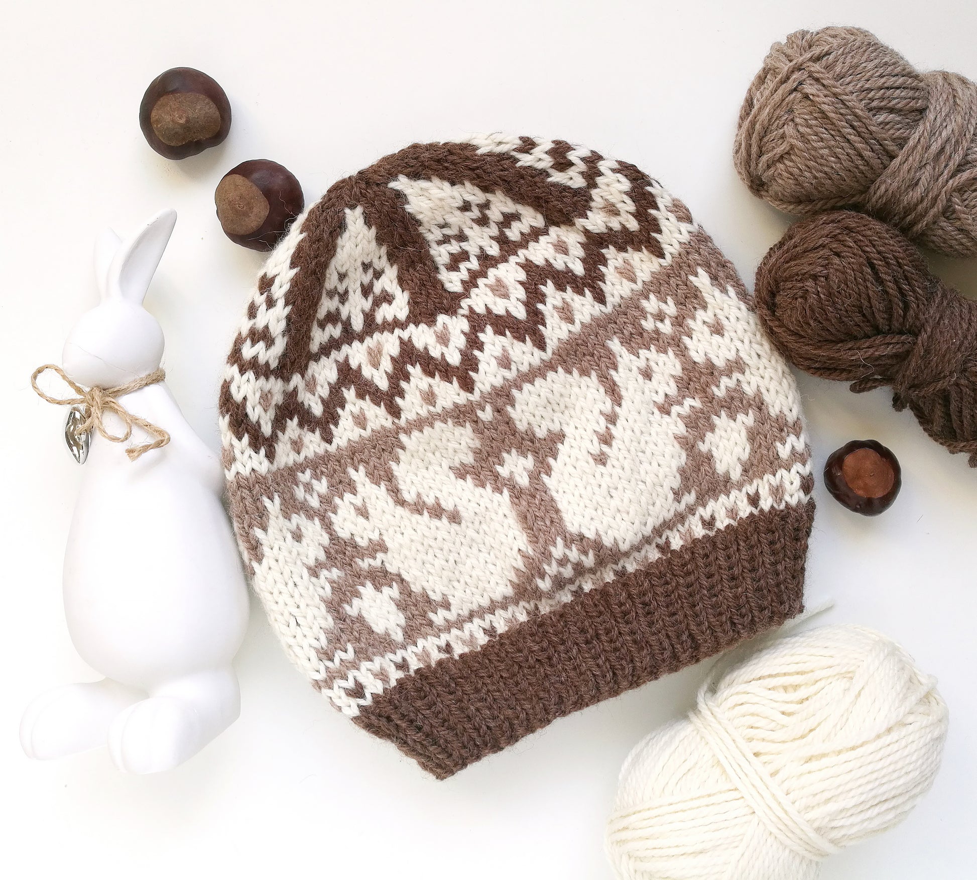 Brown and white hand-knitted Fair Isle beanie hat with a squirrel motif styled in a flat lay with yarn balls, chestnuts, a white ceramic rabbit figurine.