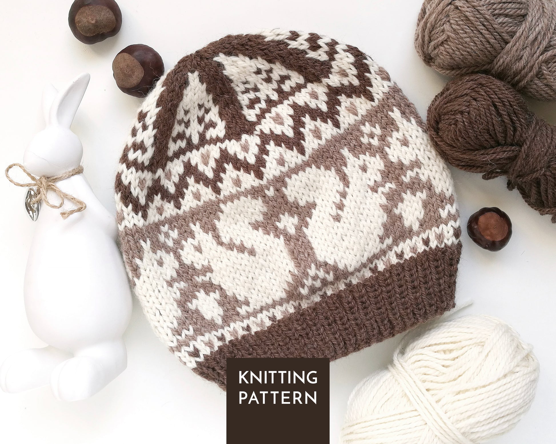 A knitted beanie hat with a brown and cream Squirrel Fair Isle pattern, styled in a flat lay with yarn balls, chestnuts, a white ceramic rabbit figurine.