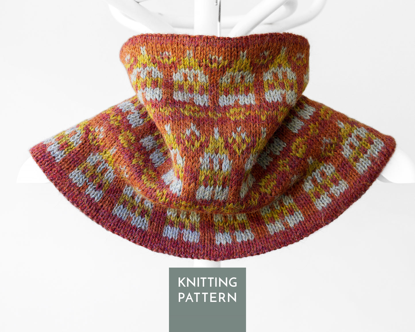 A knitted cowl neck warmer, shown draped on a white hanger against a light gray background. The knit pattern features a warm rust red base with green and light teal Fair Isle style motifs.