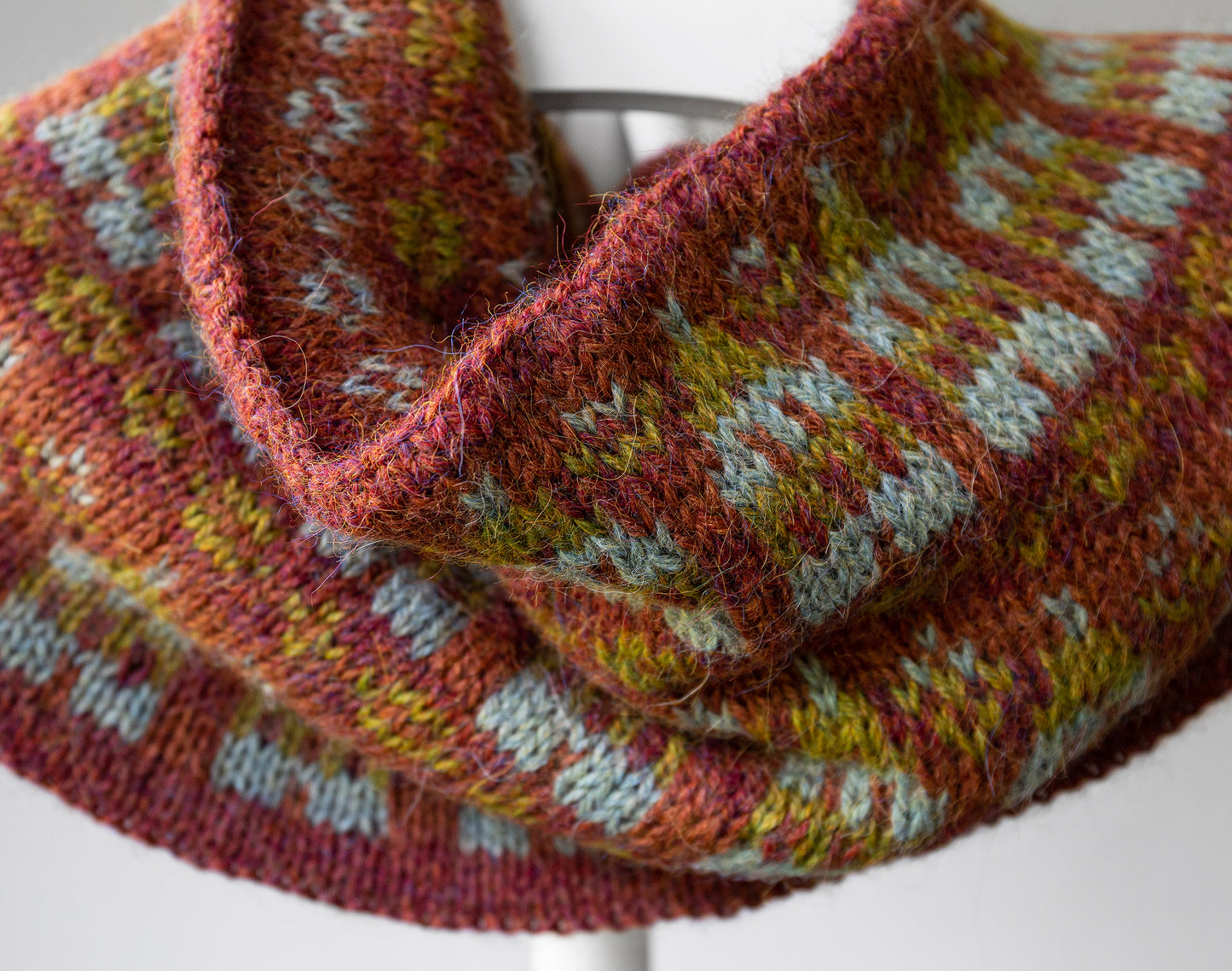 A close-up of a hand-knit cowl scarf with a multicolor fair isle pattern in rust red, mustard yellow, and pale blue