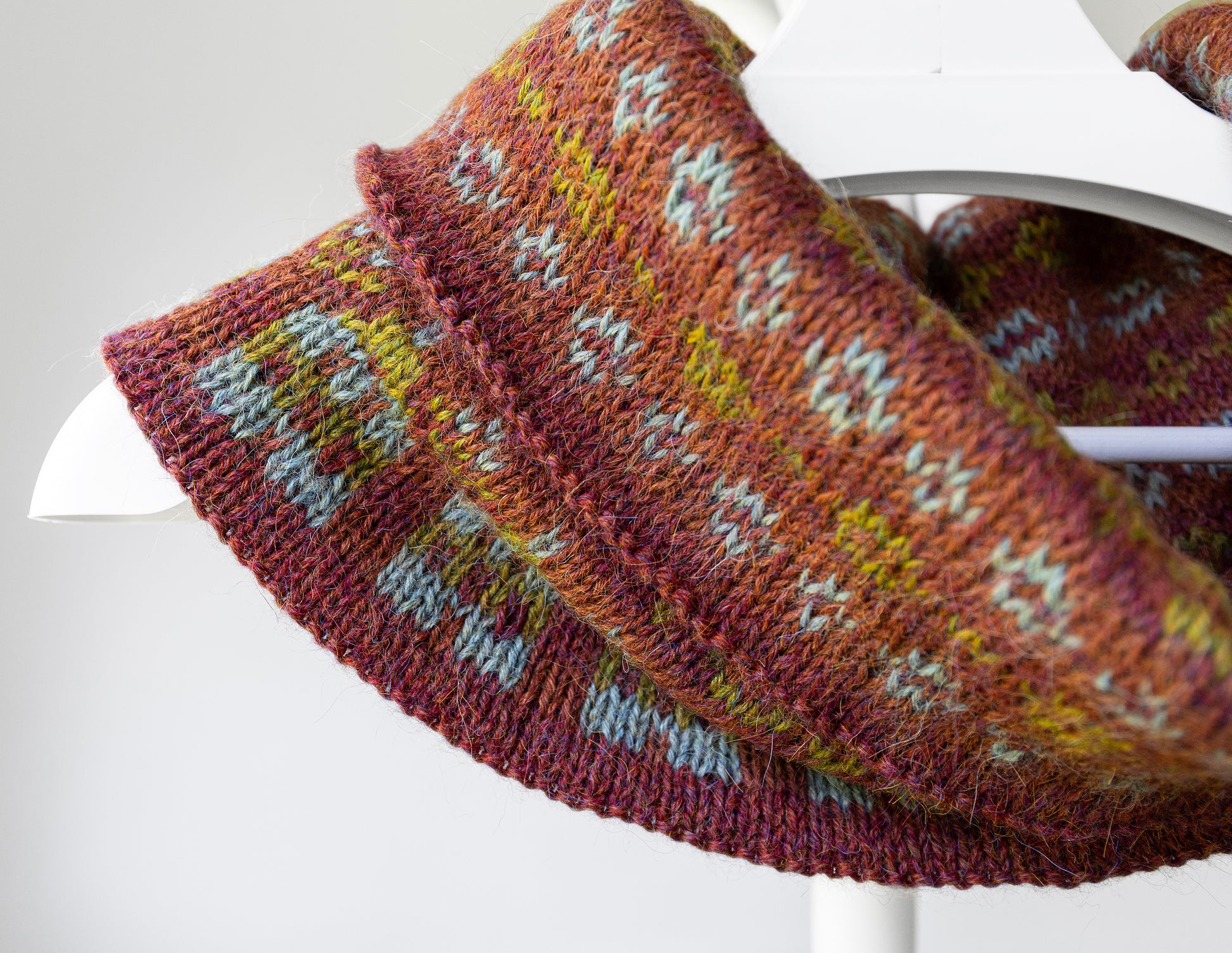 A close-up of a knitted cowl or scarf draped over a white hanger, featuring a multicolor Fair Isle pattern with rust orange, burgundy, yellow, and light blue yarn.