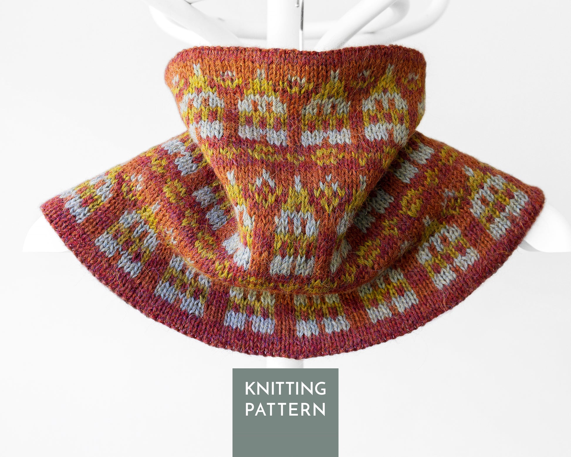 A knitted cowl or neck warmer with a wide brim, shown draped on a white hanger against a light gray background. The knit pattern features a warm rust red base with yellow and white Fair Isle style motifs.
