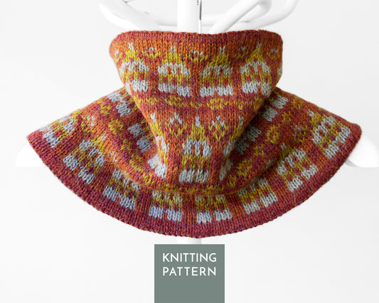 A knitted cowl or neck warmer with a wide brim, shown draped on a white hanger against a light gray background. The knit pattern features a warm rust red base with yellow and white Fair Isle style motifs.