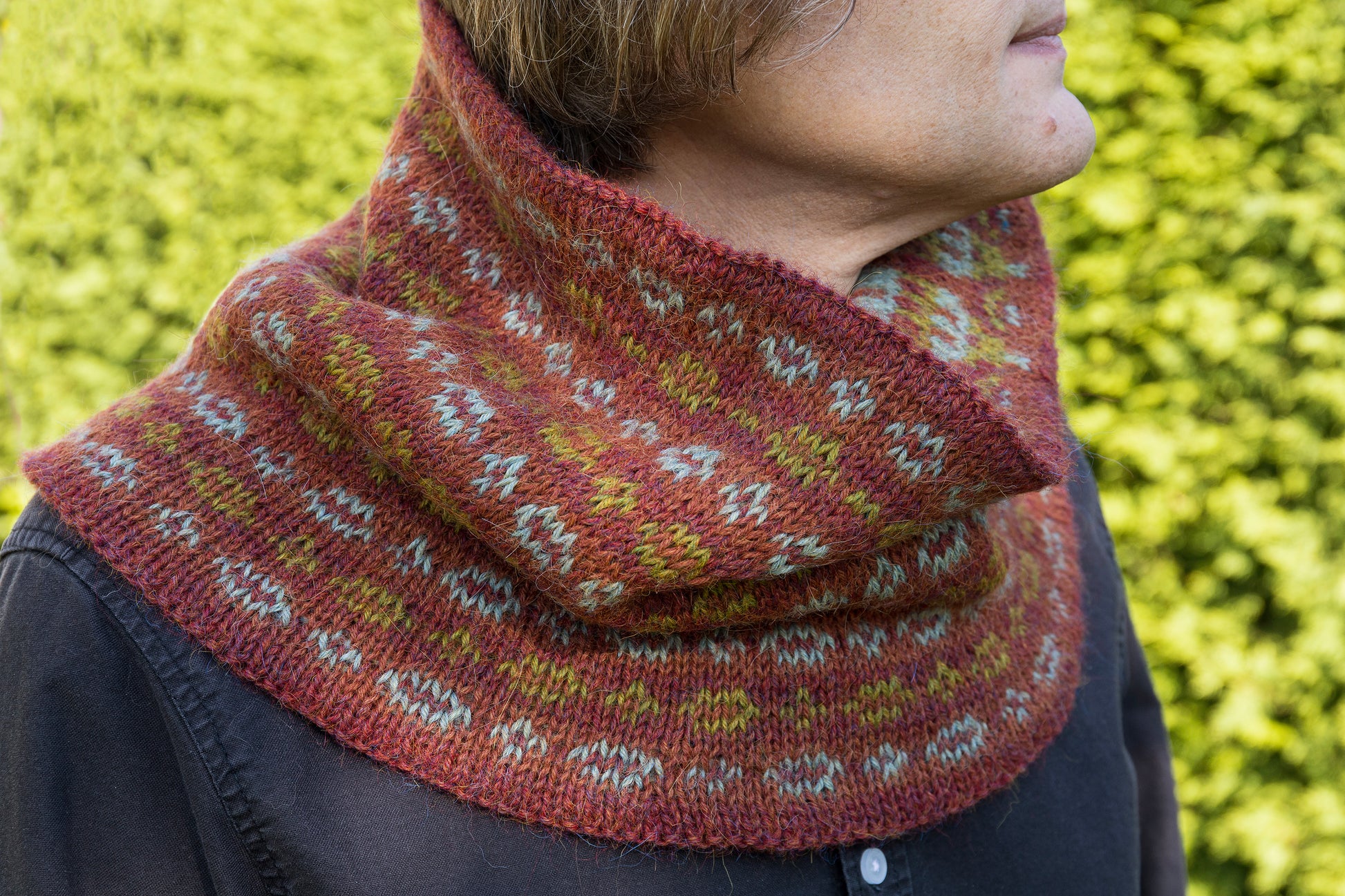 Person wearing a patterned knitted cowl against a blurred green background